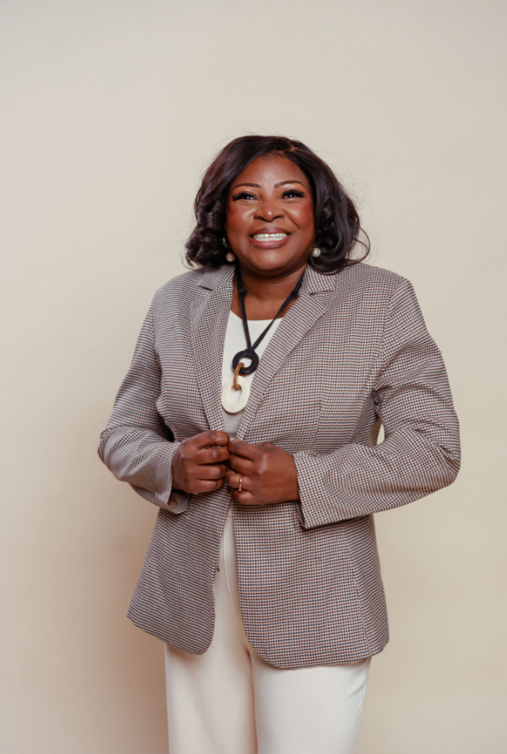 A smiling woman with dark hair, wearing a checked blazer, white top, and cream pants, standing against a plain beige background.