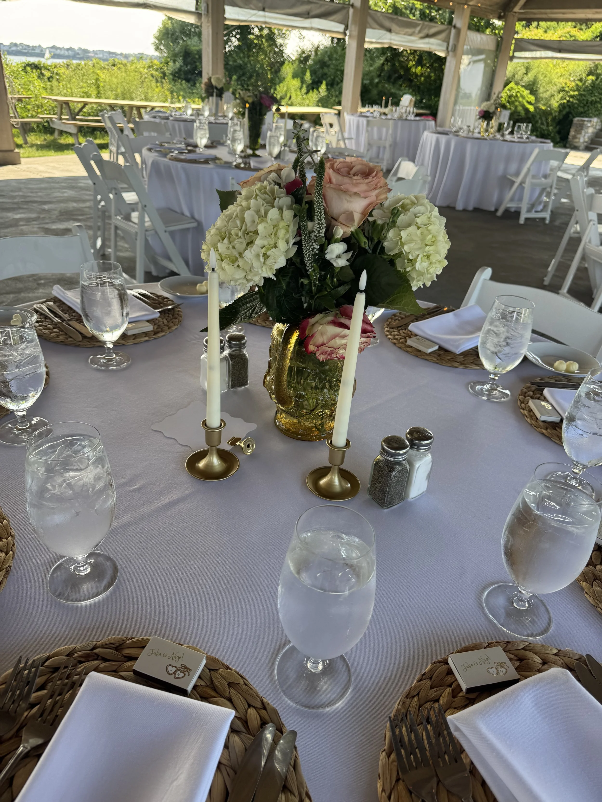 Wedding reception table set outdoors with white tablecloths, a floral centerpiece with pink and white roses, hydrangeas, and greenery, candles, and place settings with glassware and cutlery. There are white chairs and a scenic view of trees and water