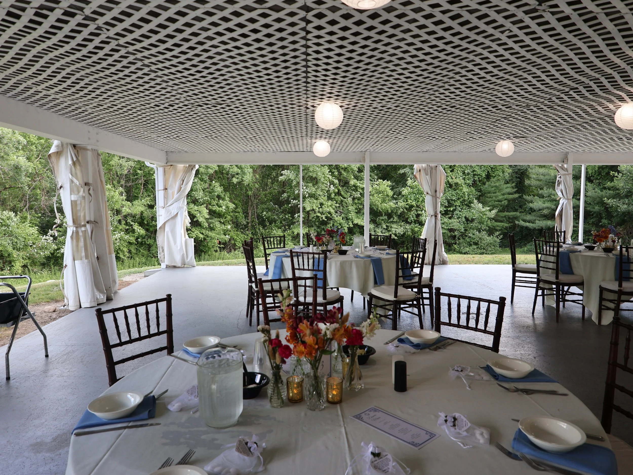 Outdoor party setup with round tables, chairs, floral centerpiece, candles, and place settings under a decorated canopy with string lights, surrounded by green trees.