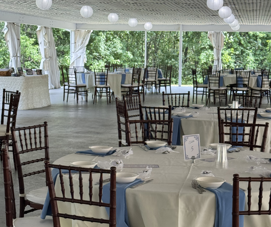 Indoor event space decorated with round tables covered with white tablecloths and blue runners, set with bowls, plates, and utensils, surrounded by dark brown wooden chairs, with large windows and white paper lanterns hanging from the ceiling, overlo