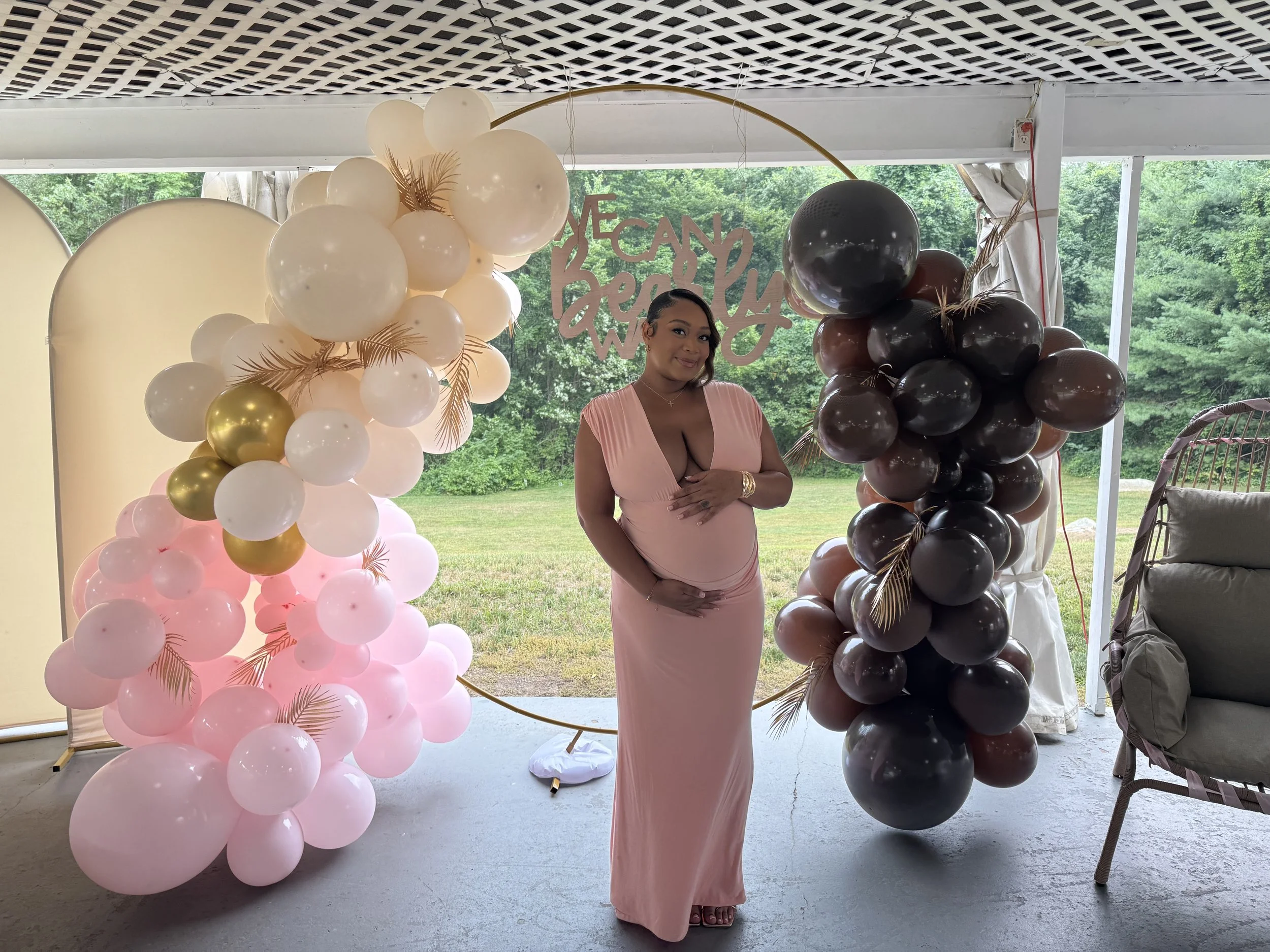 A woman in a pink dress standing in front of a large balloon display with pink, white, gold, and black balloons at an outdoor event.