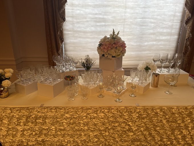 Elegant table display with various glassware, floral arrangements, and a gold-colored tablecloth, set near a window with curtains.