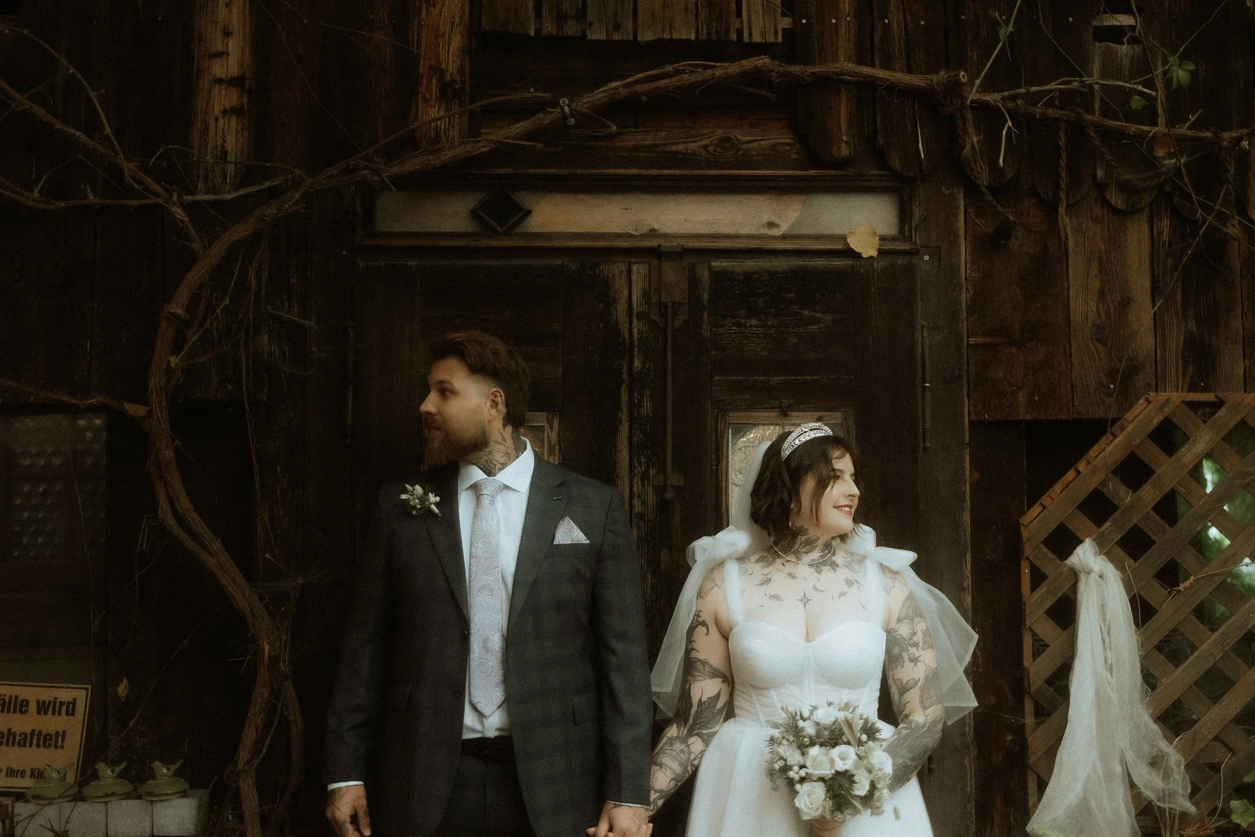 A bride and groom holding hands, standing side by side in front of a rustic wooden backdrop. The bride has tattoos and is holding a bouquet, smiling, while the groom is looking at her.