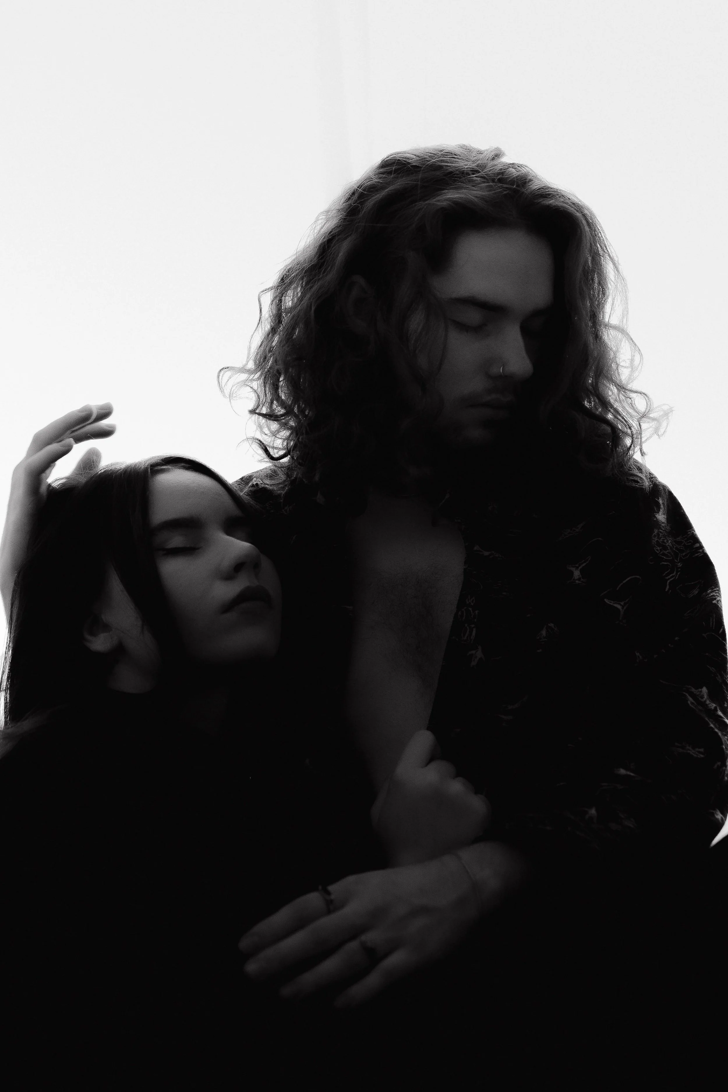 Black and white photograph of a man and woman embracing, with eyes closed, their faces peaceful. The image has a minimalist background and artistic lighting.