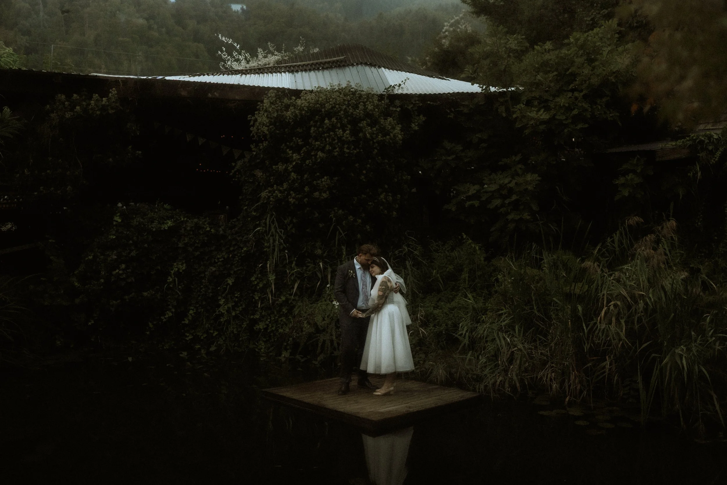 A couple in wedding attire standing close on a small wooden platform by a pond, surrounded by dense greenery, with a rustic building with a metal roof in the background.