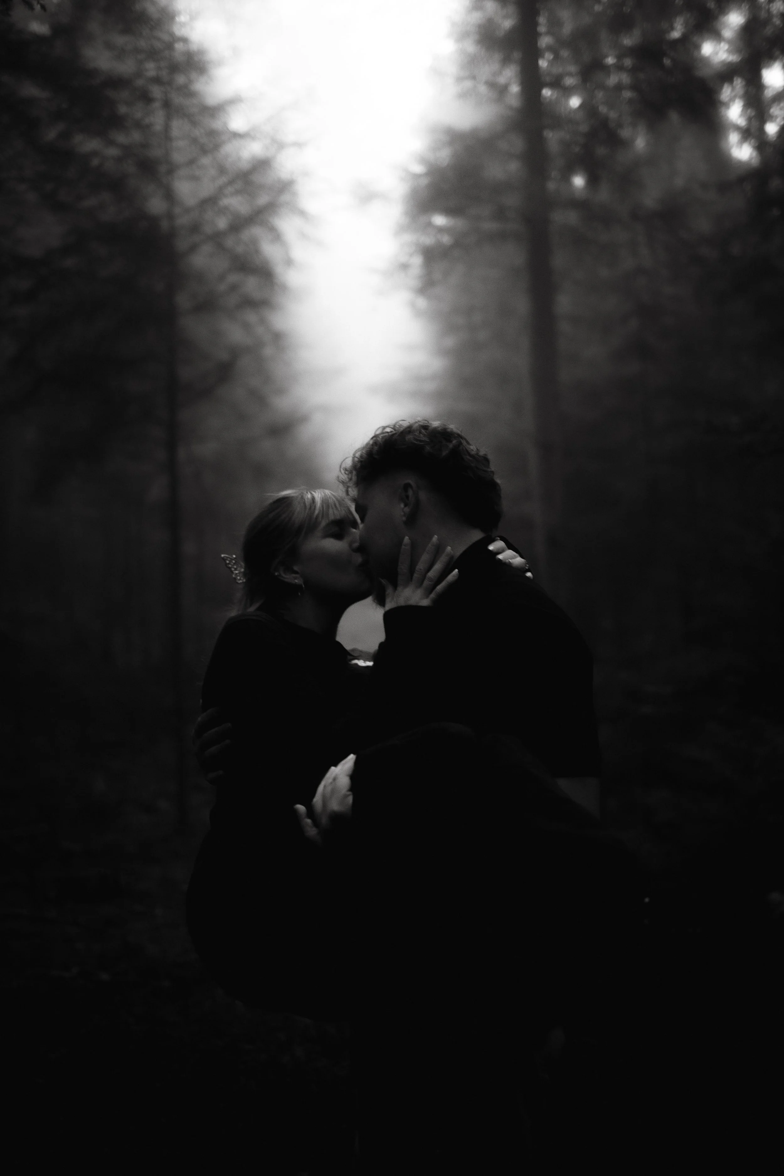 A black and white photo of a couple kissing in a forest at sunset, with tall trees and fog in the background.