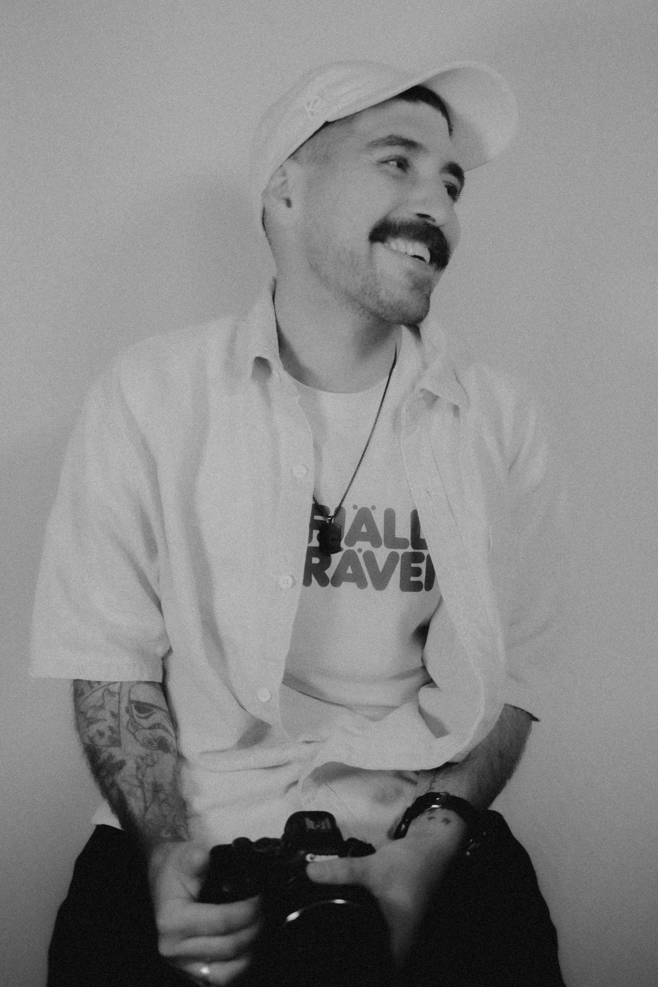 A smiling man with a mustache and tattoos, wearing a cap, a graphic t-shirt, and an open button-up shirt, holding a camera, sitting against a plain background.