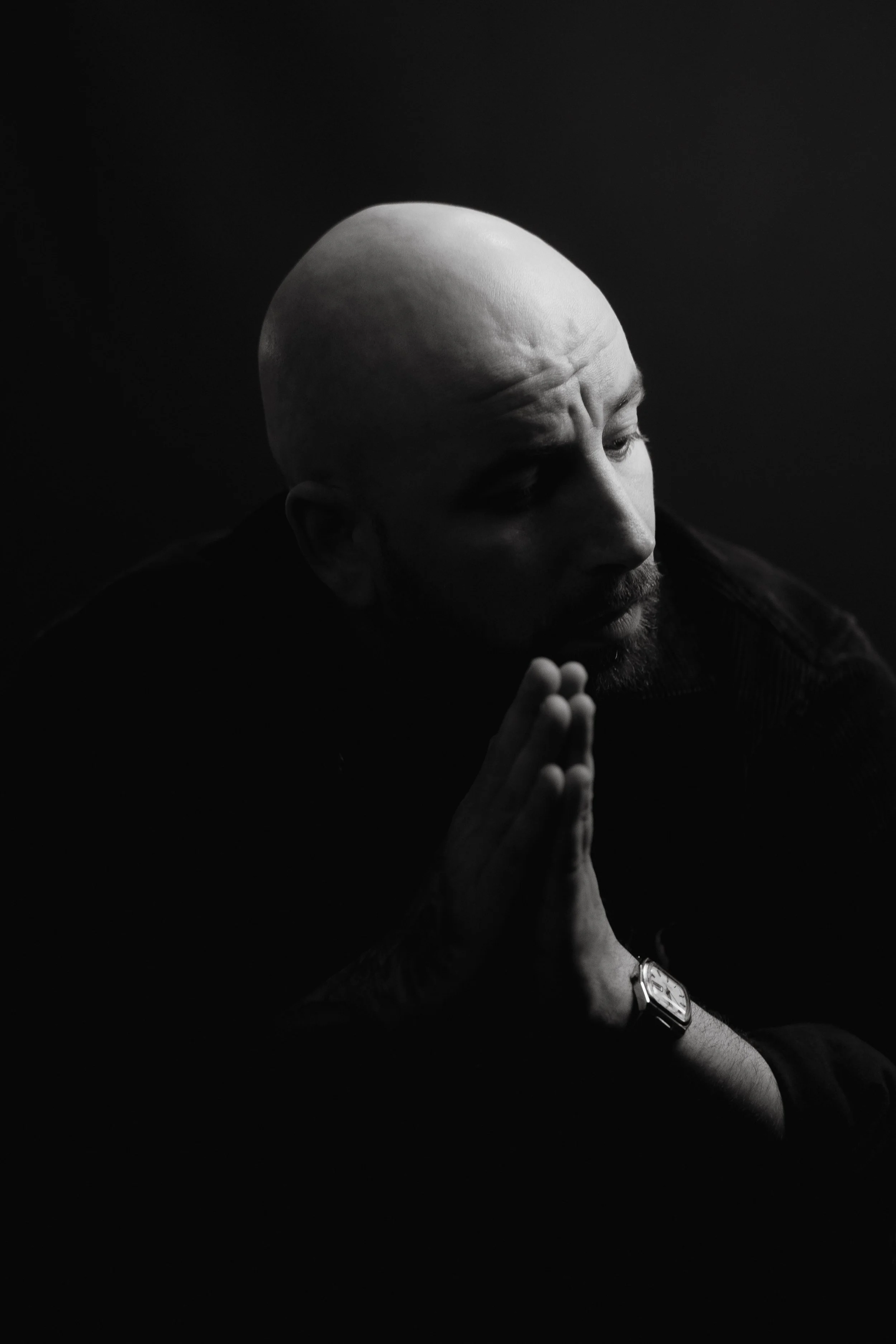 Black and white photograph of a man with a shaved head and beard, holding his hands together in a prayer or meditation pose, wearing a wristwatch, against a dark background.