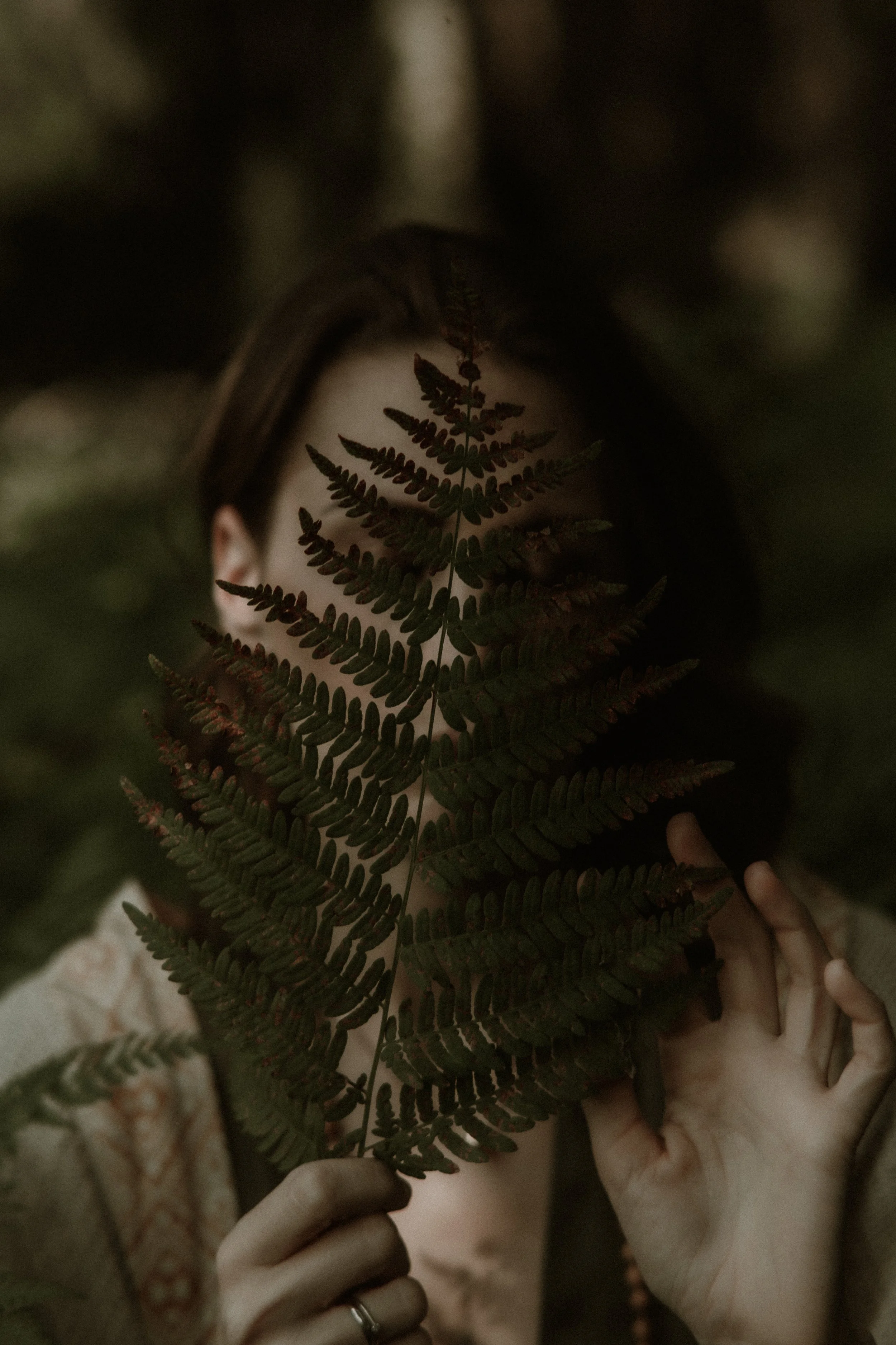 A person holding a large fern leaf in front of their face, partially obscuring their facial features. The person has light skin and dark hair, and the background is blurred with a natural, forest-like setting.