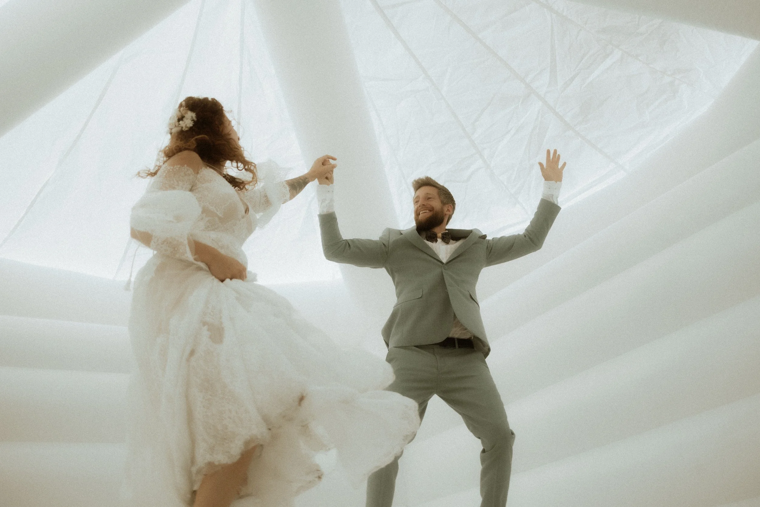 A bride and groom dancing inside a modern, white, tent-like structure with translucent ceiling panels, with the groom smiling and the bride in a lace wedding dress.