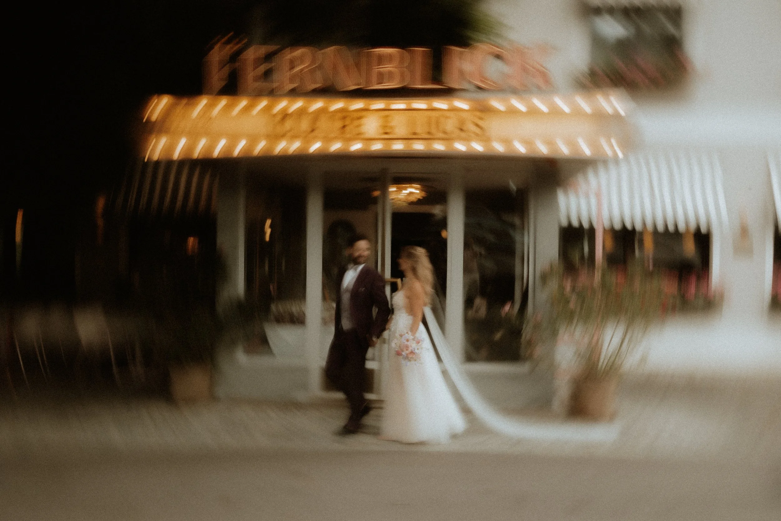 A bride and groom holding hands outside a venue at night with a brightly lit sign above them.