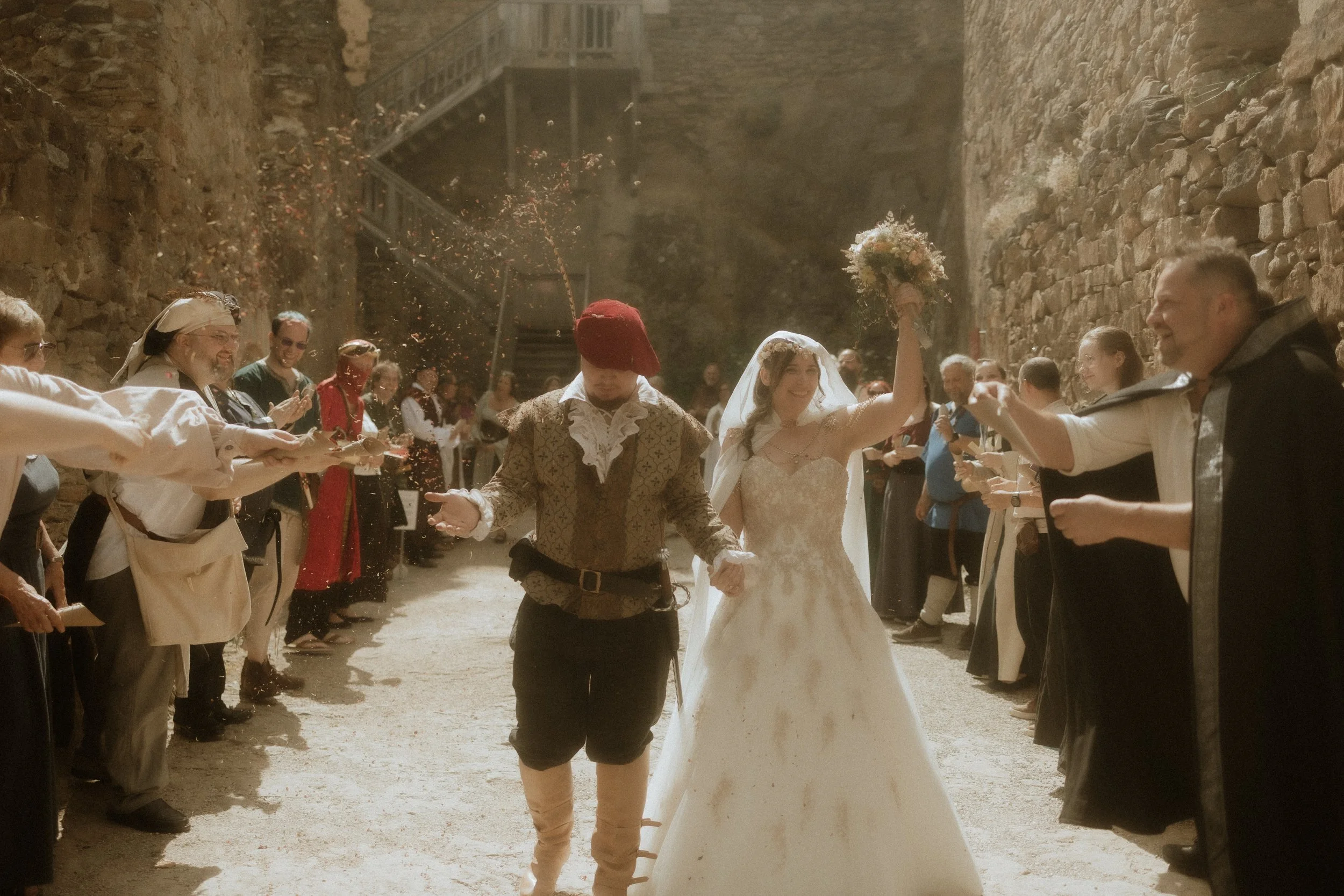 Wedding celebration with bride and groom walking through an arch of guests outside, surrounded by stone walls, guests throwing confetti, and the bride holding a bouquet, smiling, with guests clapping and celebrating.