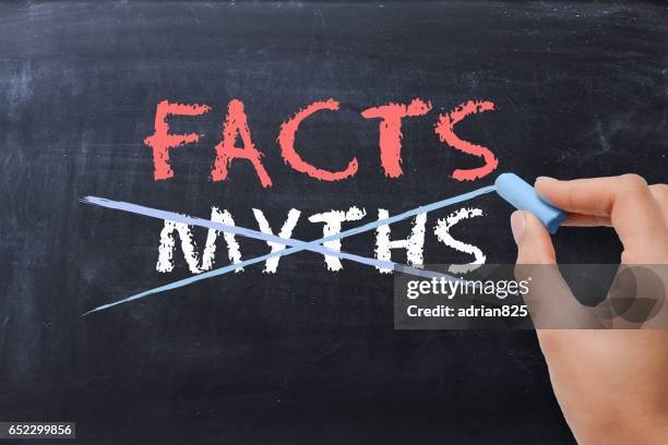 A person erasing the word 'MYTHS' from a chalkboard, leaving the word 'FACTS' visible in red, with a blue cloth or eraser in hand.