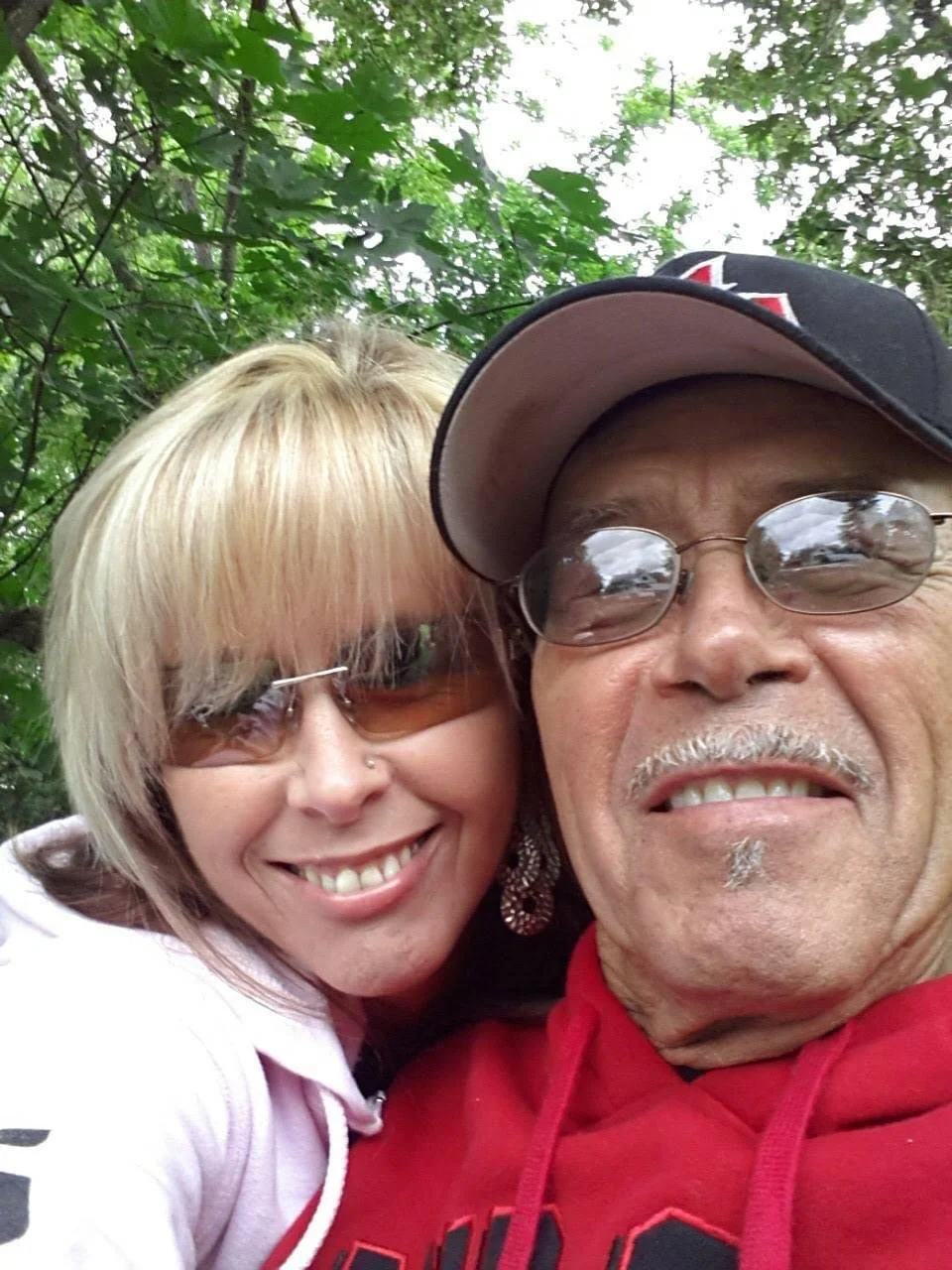 Close-up of a smiling woman with blonde hair and sunglasses, and an older man wearing a baseball cap, sunglasses, and a red hoodie, outdoors with green foliage in the background.