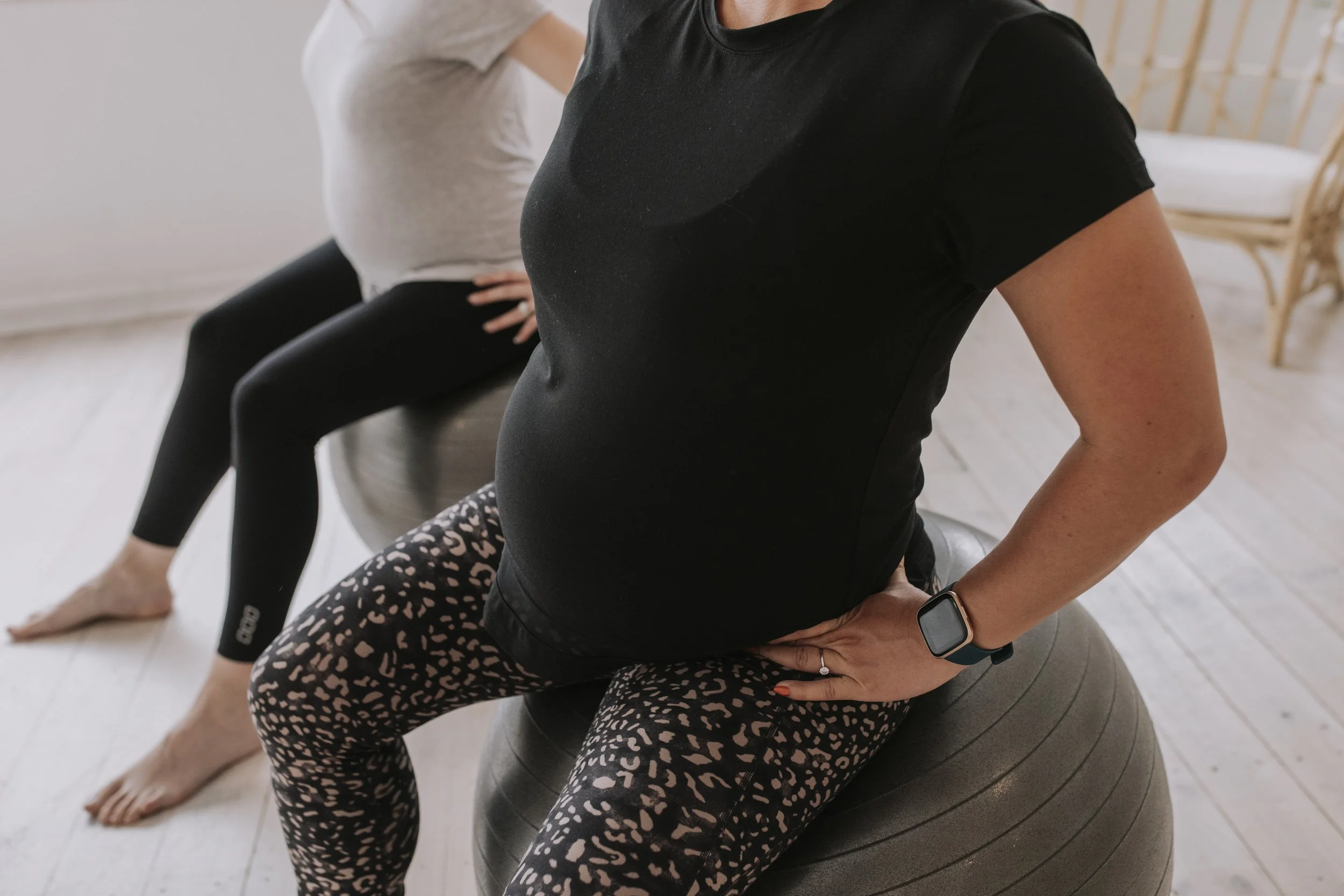 A pregnant woman sitting on an exercise ball during a fitness or prenatal exercise class, with another person sitting behind her.