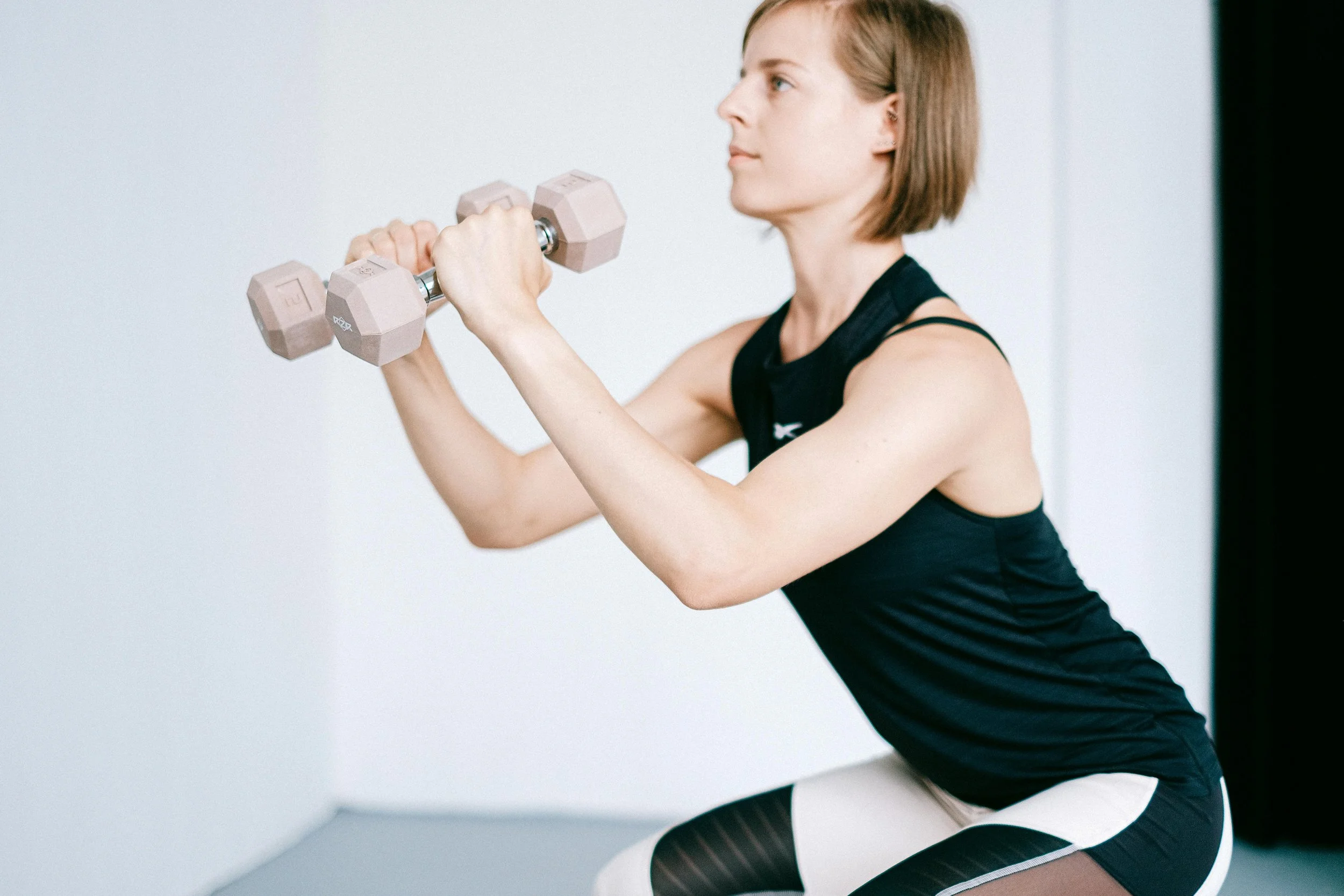 A woman in workout attire performing a squat exercise with light pink dumbbells in a gym or fitness studio.