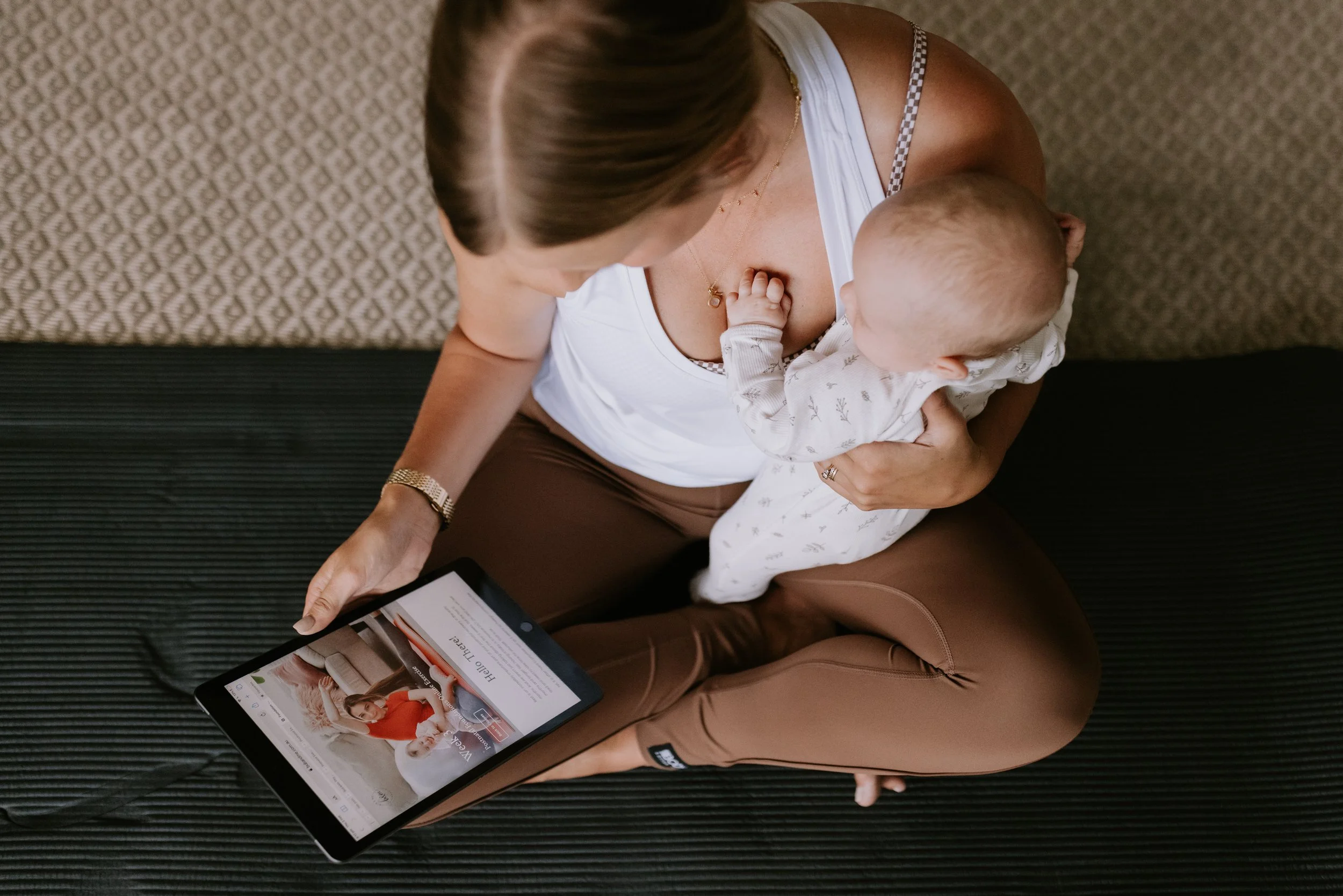 A woman sitting cross-legged on a black mat, breastfeeding a baby while looking at a tablet, with a textured beige wall behind her.