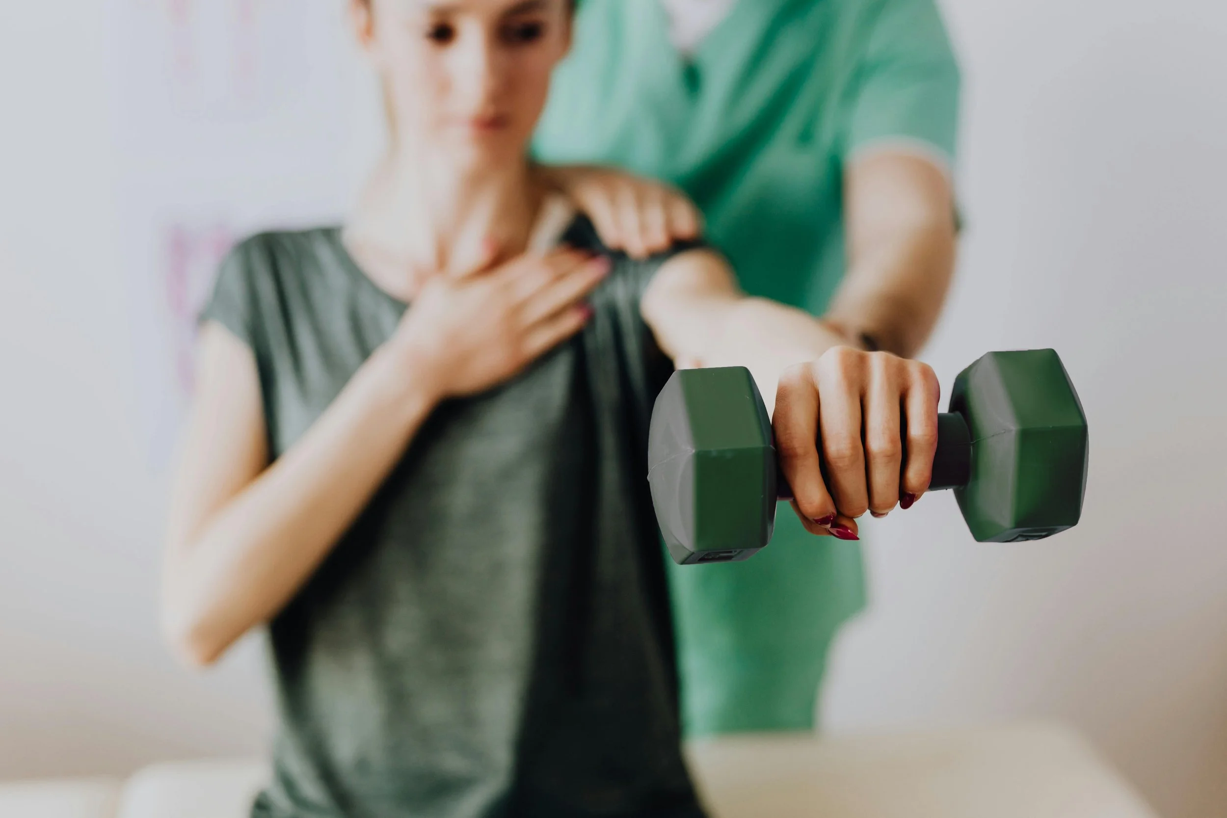 A woman undergoing physical therapy, with a therapist assisting, as she lifts a green dumbbell in a therapy session.