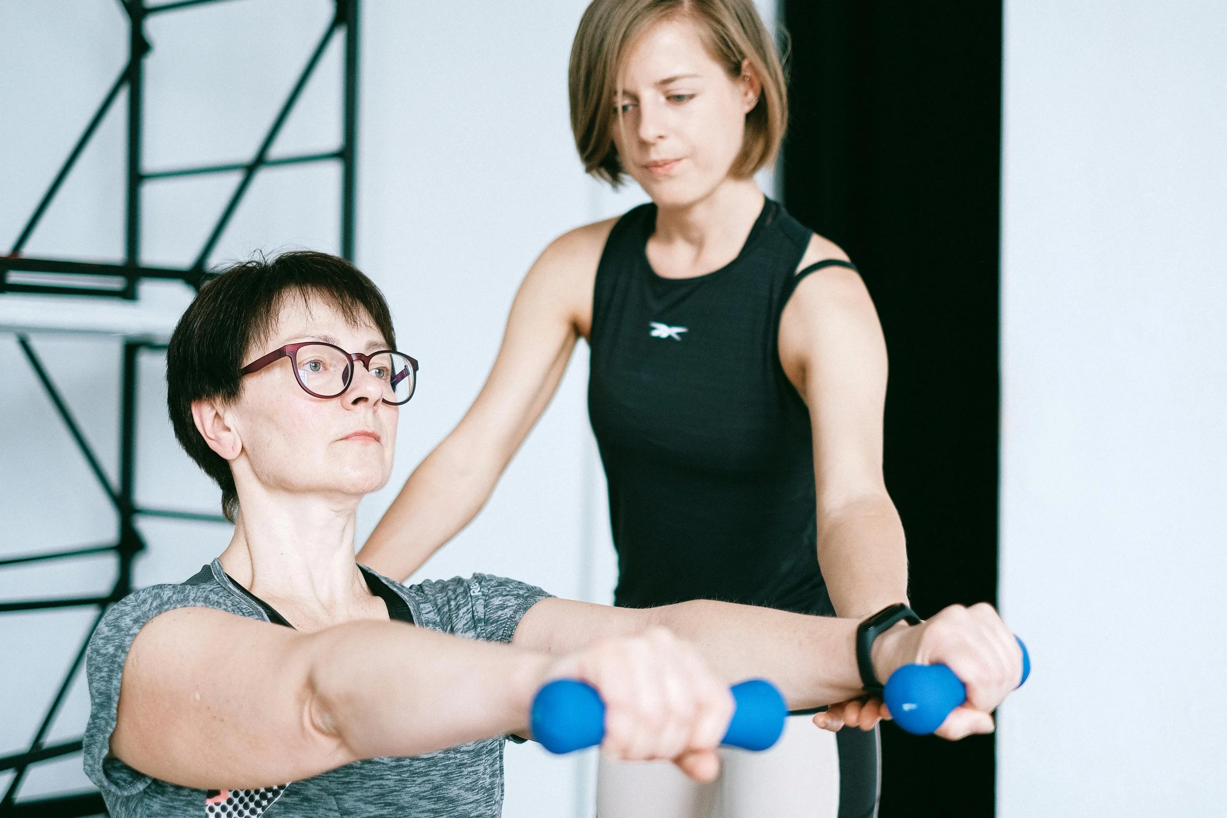 A woman assisting a person with a disability during a gym workout, guiding their arm as they lift blue dumbbells.