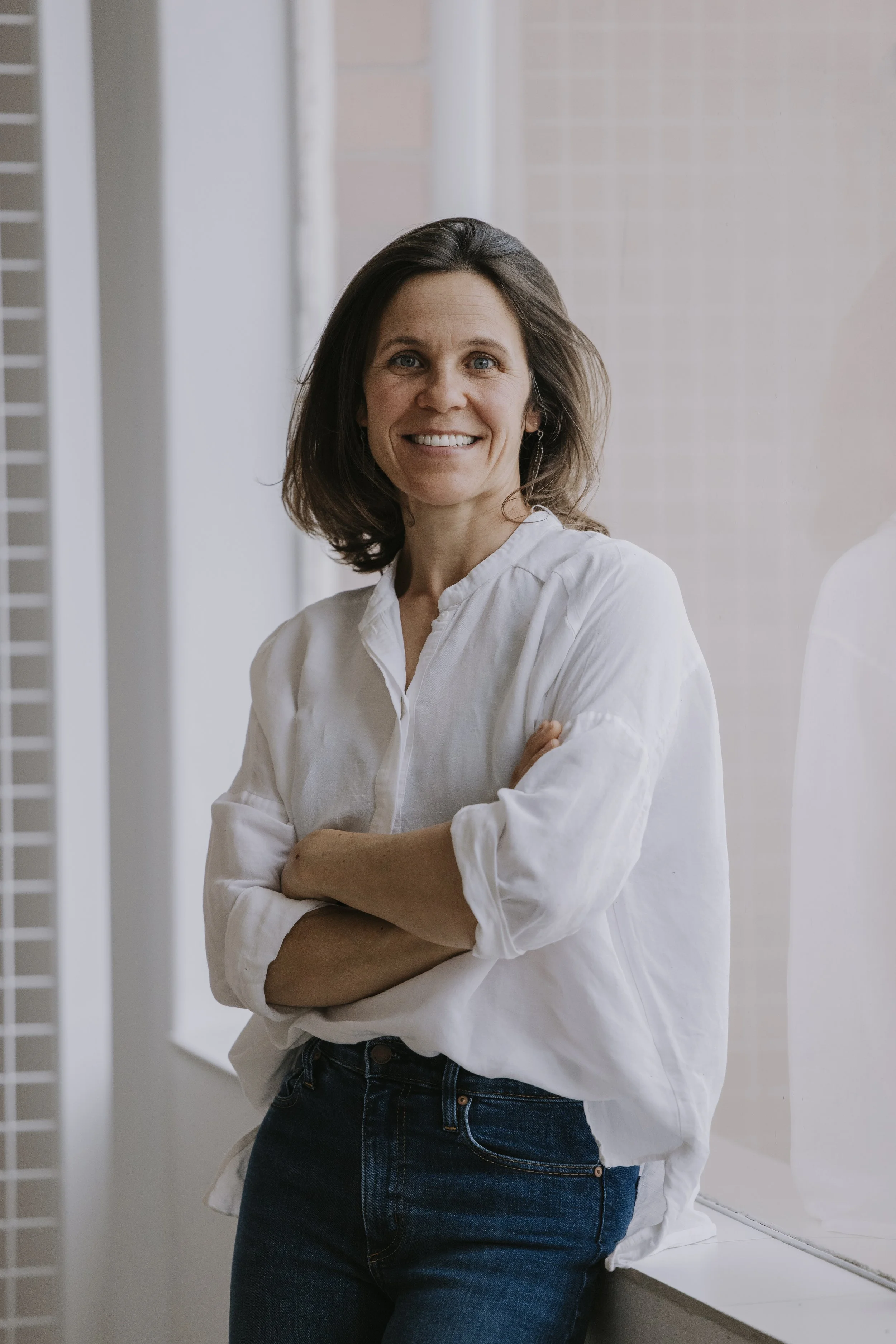 A smiling woman with shoulder-length brown hair, wearing a white blouse and dark jeans, standing with arms crossed indoors near a window.