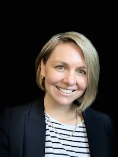 A woman with short blonde hair smiling, wearing a black blazer over a striped shirt, against a black background.