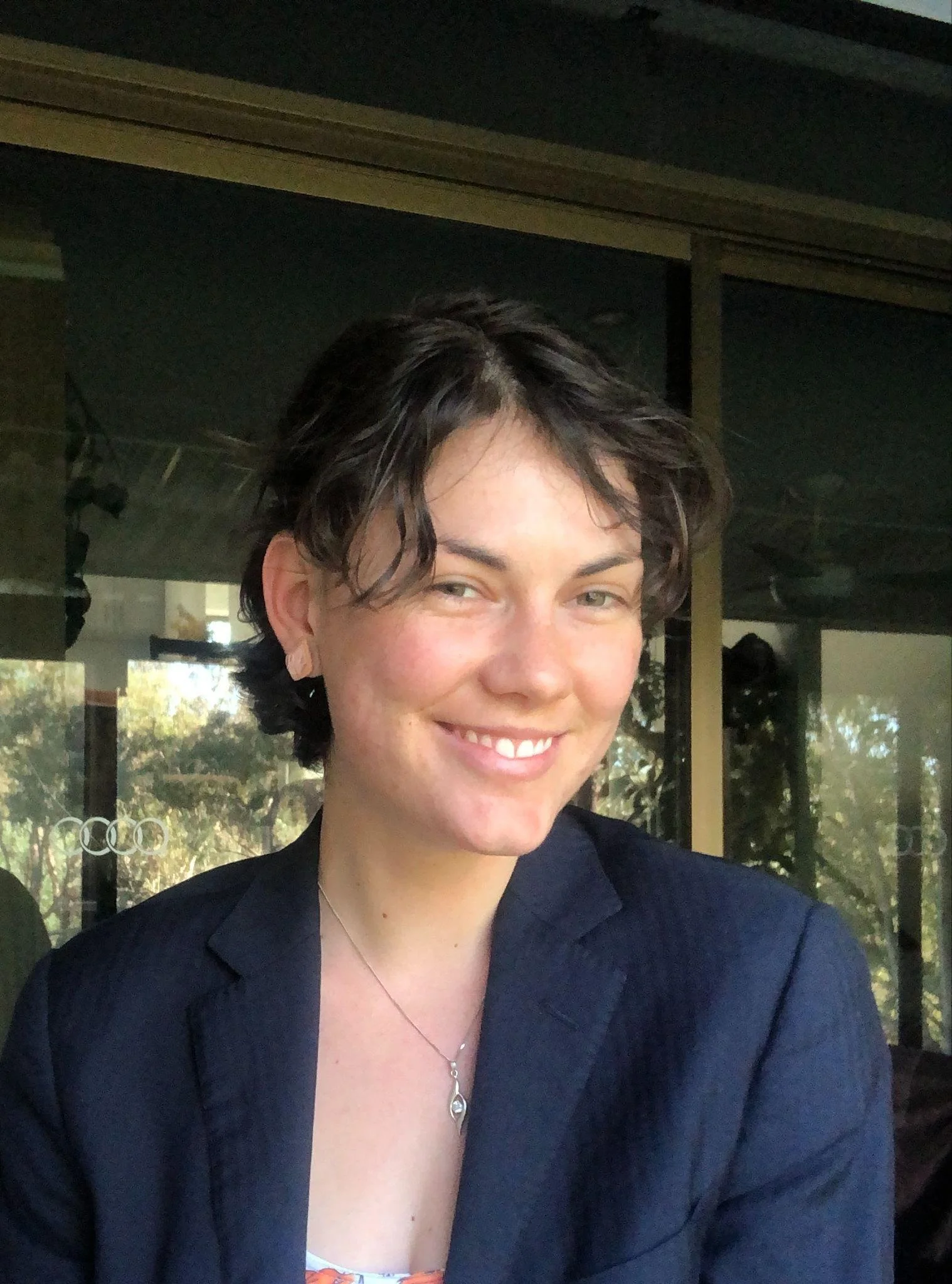 A young woman with short, wavy dark hair and fair skin, smiling, wearing a black blazer and a necklace, sitting indoors with large windows showing trees outside.