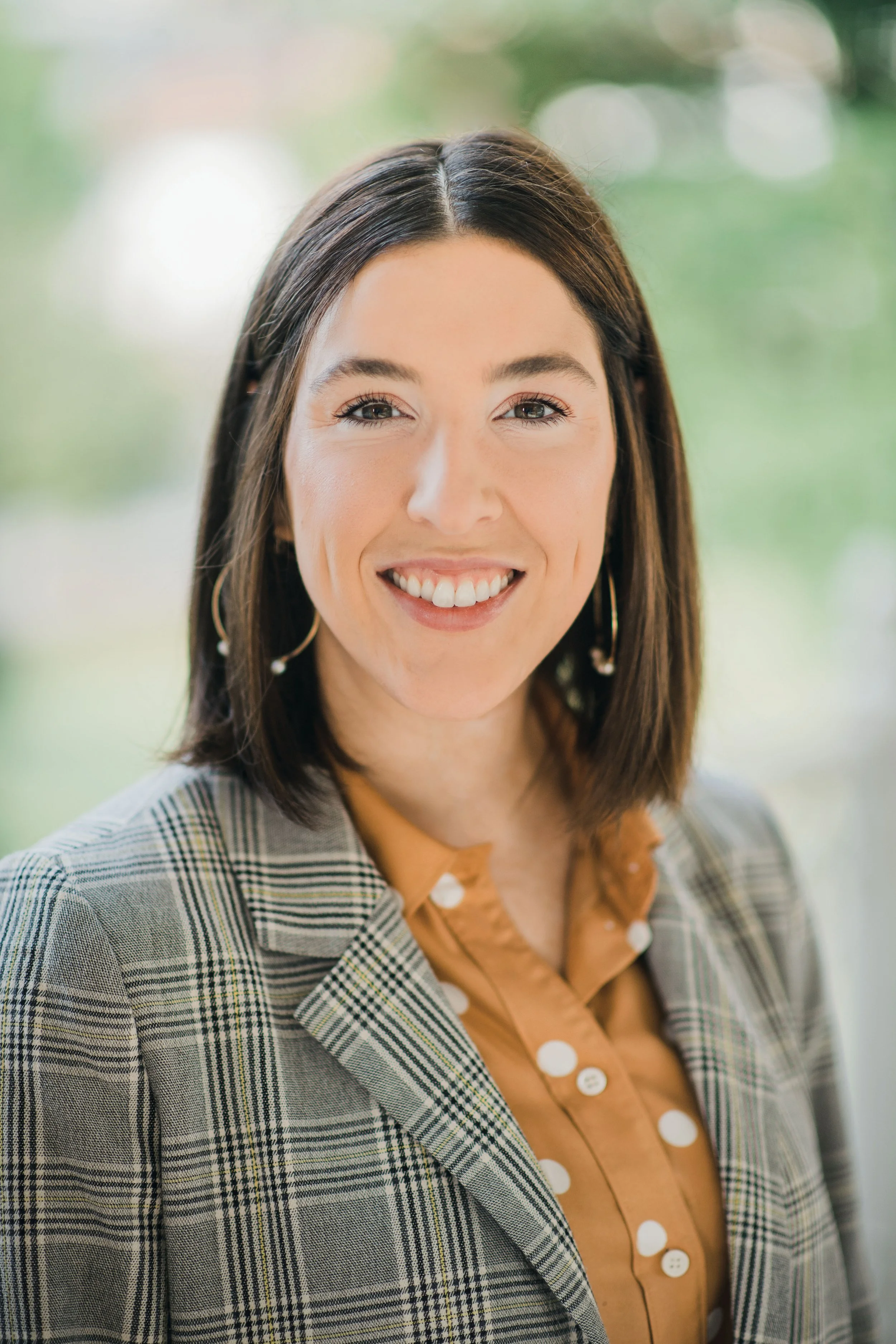 A woman with shoulder-length brown hair, wearing a plaid blazer over a brown blouse with white polka dots, smiling at the camera.