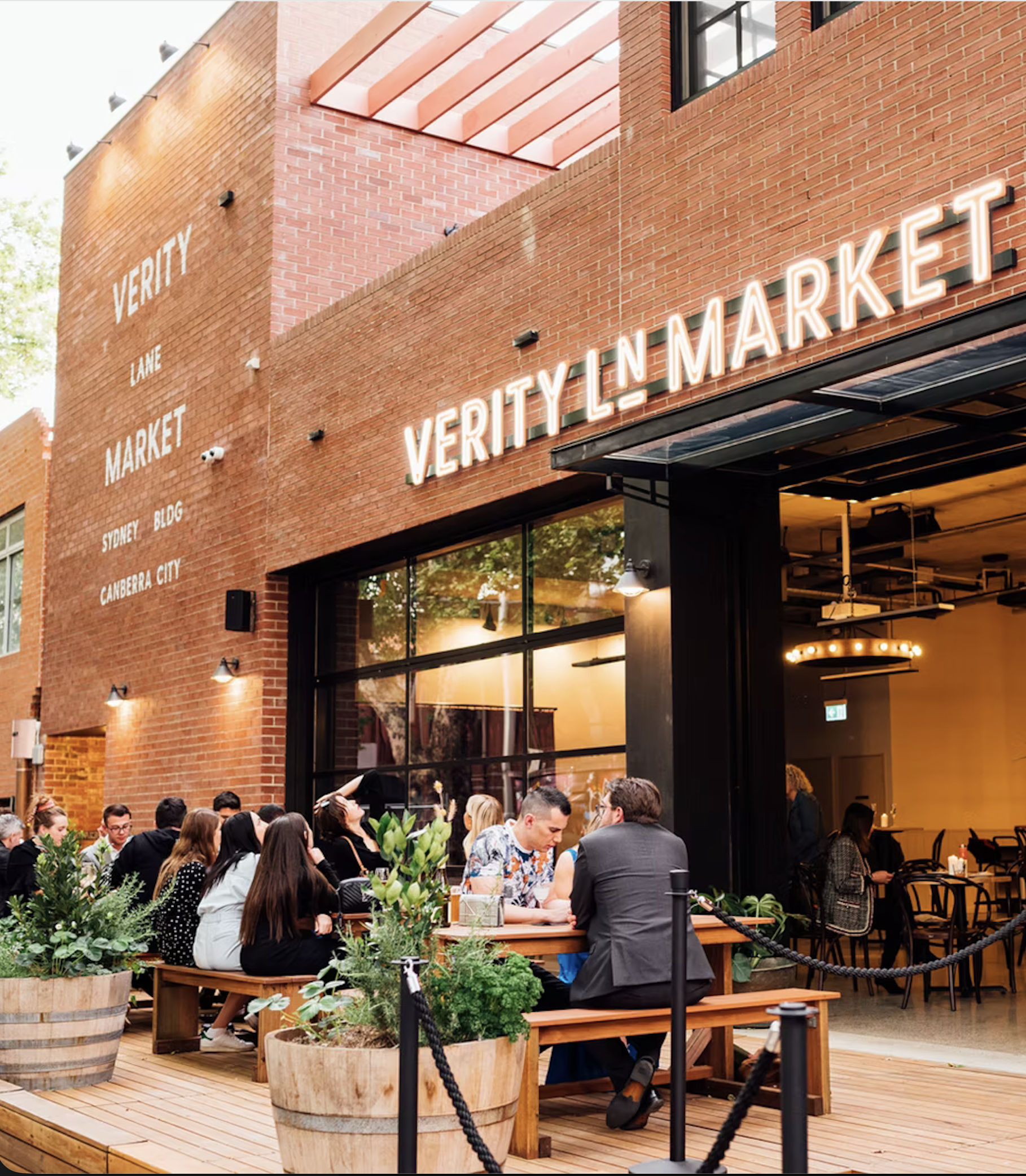 People dining outside at Verity Lane Market, a brick building with a neon sign and large glass windows, with potted plants and string lights.