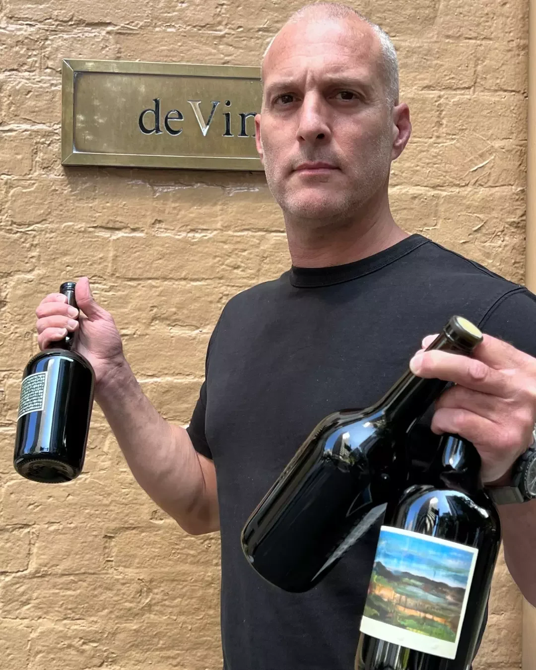 A somelier holding  two bottles of wine in front of a brick wall with a sign that reads "de Vino".