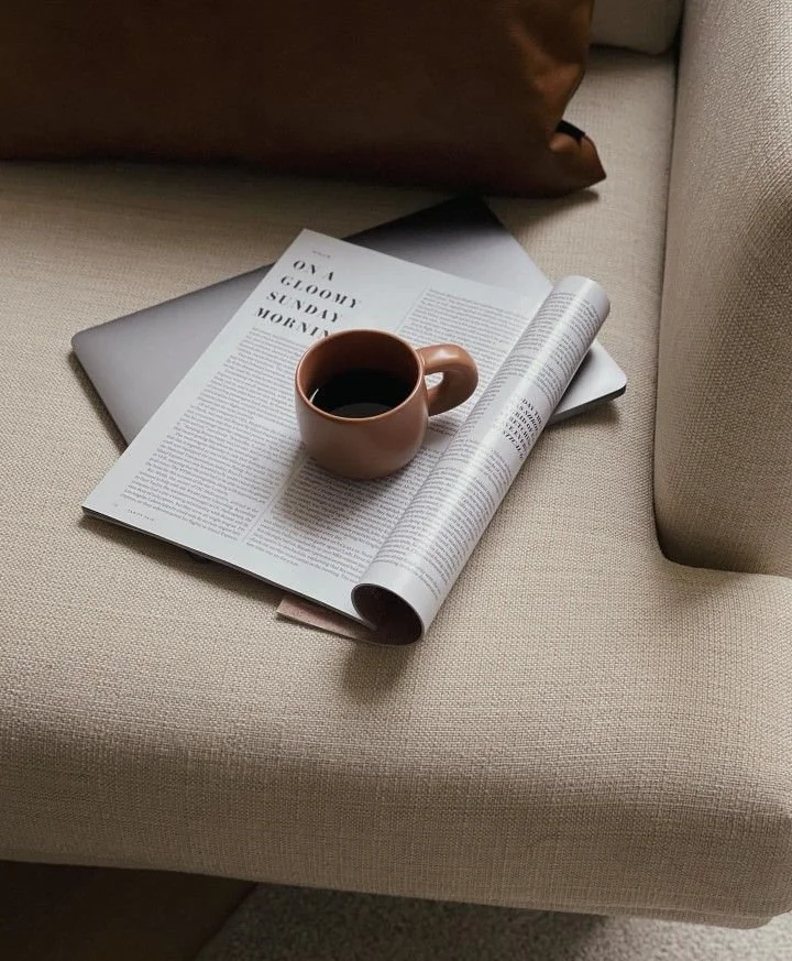 A beige couch with a mug of black coffee, an open magazine, a closed silver laptop, and a brown cushion.