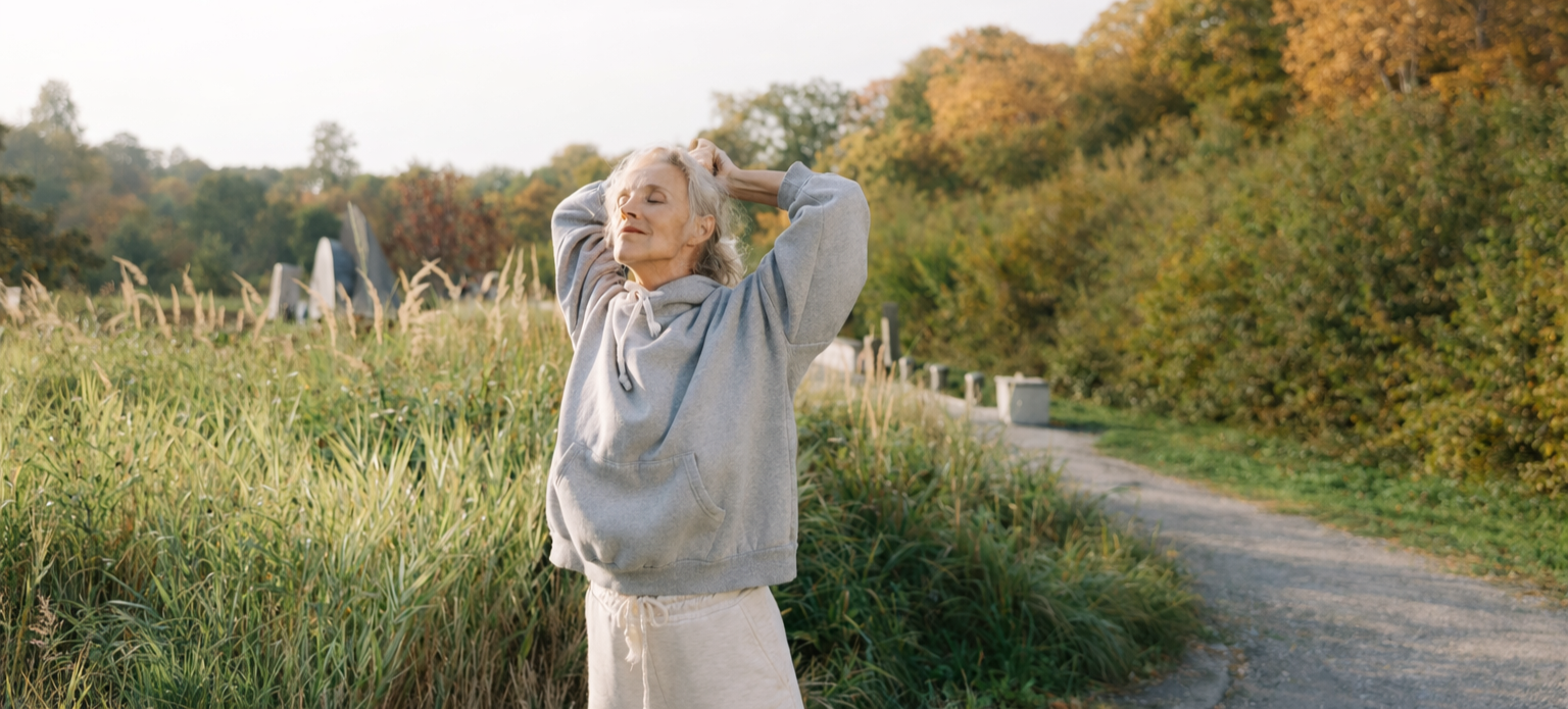 An elderly woman in a gray hoodie and beige shorts standing on a dirt path with her hands behind her head, enjoying the outdoors surrounded by trees and tall grass during autumn.