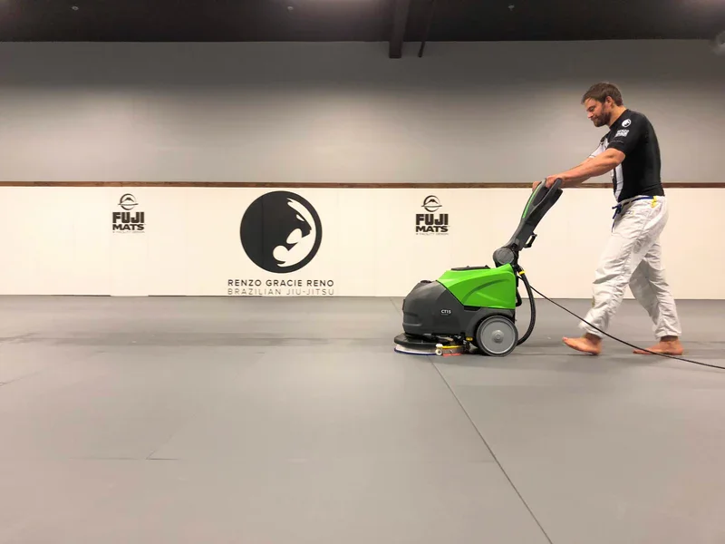 A man cleaning a martial arts training mat with a green and black floor cleaning machine at Renzo Gracie Reno Brazilian Jiu-Jitsu gym.
