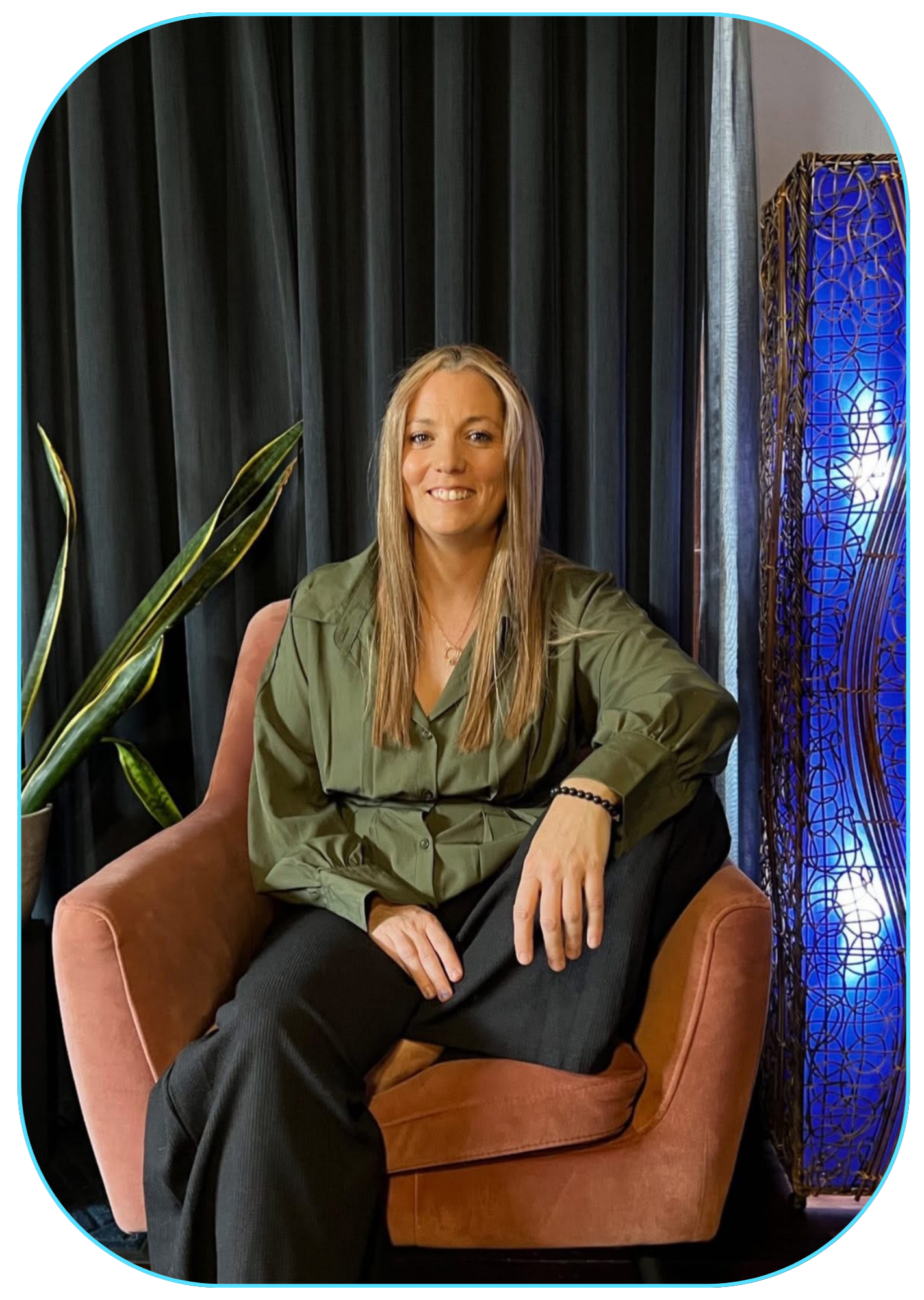 A woman with long blonde hair, wearing a green shirt and black pants, seated in a pink velvet armchair with a plant and dark curtain behind her, and a blue decorative light fixture on her right.