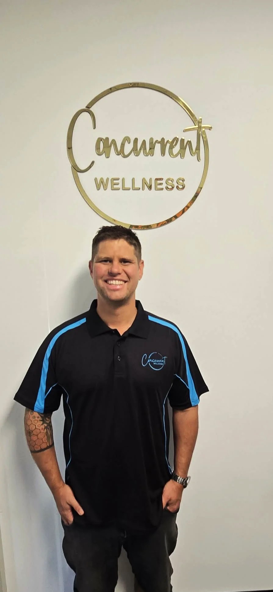 A smiling man standing against a white wall with a gold circular sign that reads 'Current Wellness' and a logo on his shirt matching the sign.