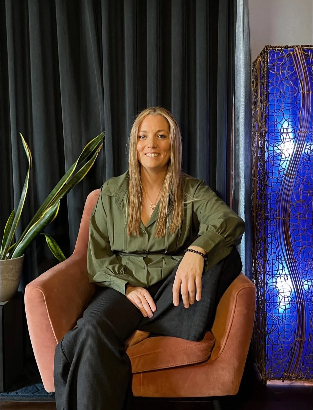 A woman with long blonde hair sitting on a peach-colored armchair, smiling, wearing a green blouse and black pants, with a black bracelet on her left wrist. There is a large plant to her left and a decorative blue light panel to her right, with black curtains in the background.