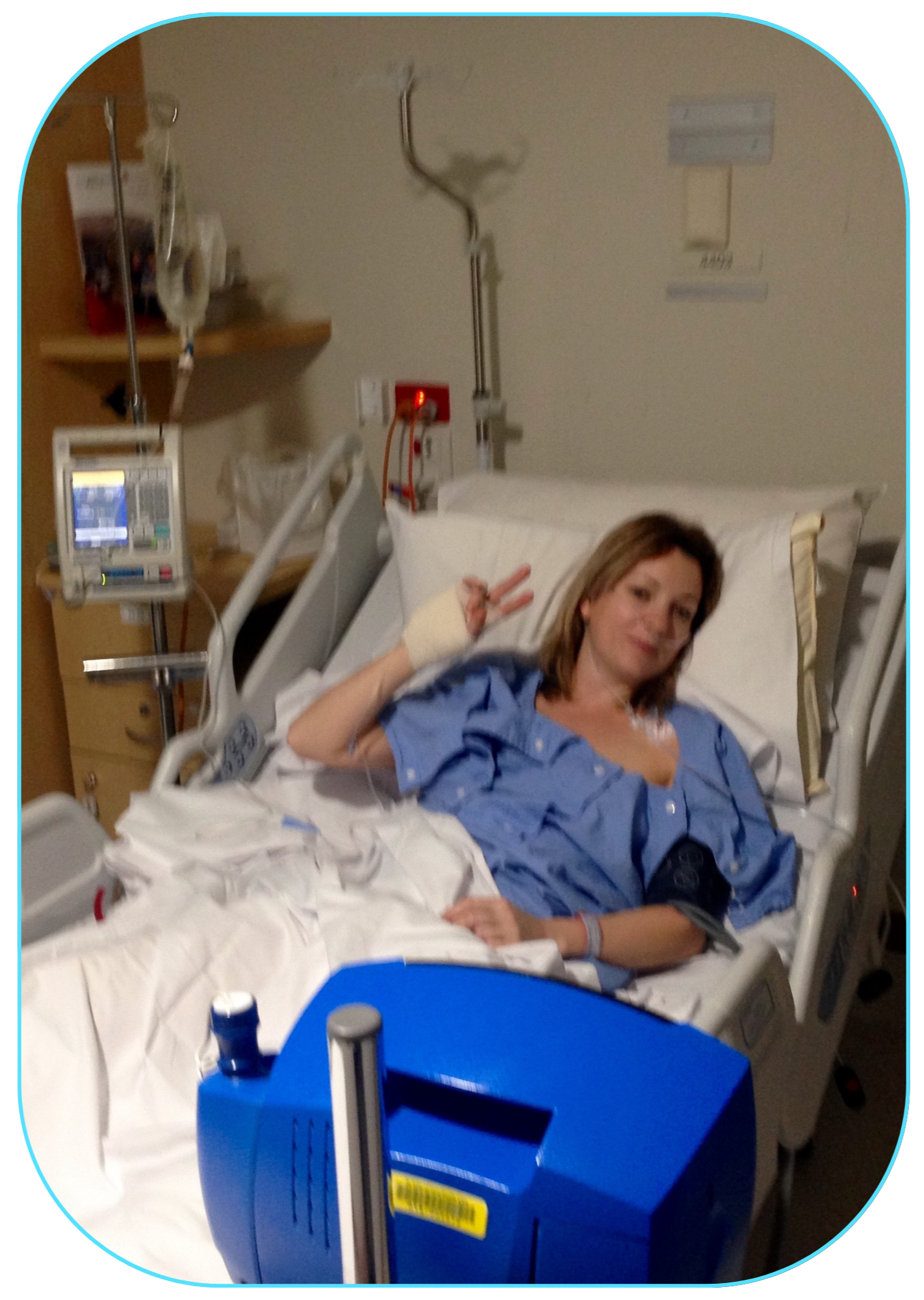 A woman lying in a hospital bed making a peace sign with her hand and smiling.