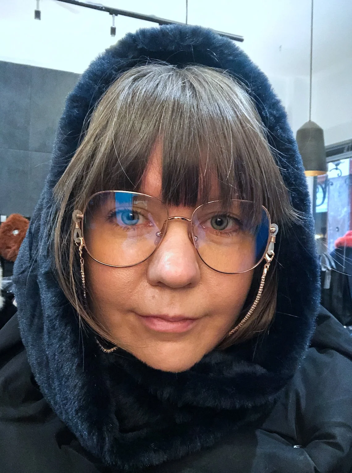 A person wearing large glasses with a gold chain, a black fluffy hood, and a black coat, seated indoors with a neutral expression.