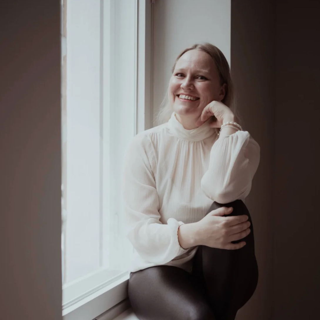 A woman with blonde hair smiling and sitting by a window, resting her chin on one hand. She wears a cream-colored blouse and dark pants.
