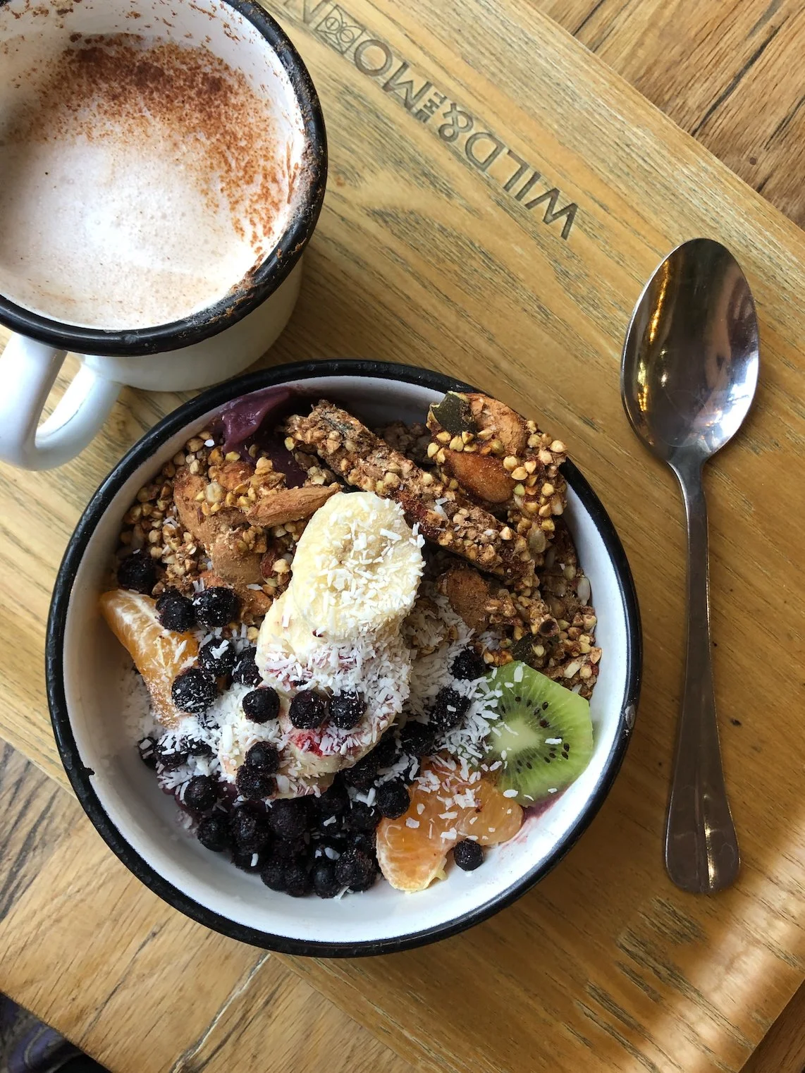 A bowl of fruit and granola topped with sliced banana, shredded coconut, blueberries, and kiwi slices, placed on a wooden table alongside a cup of frothy coffee and a spoon.