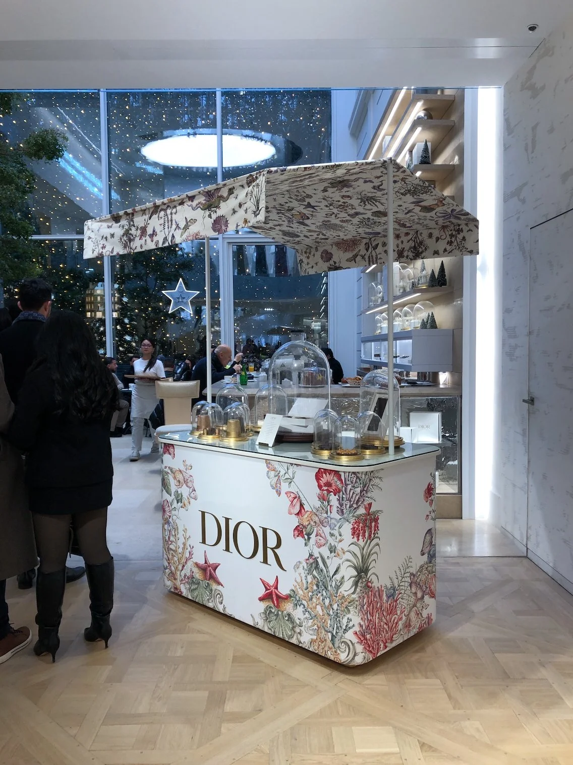 Dior pop-up shop with a floral design and glass display covers, set inside a modern, festive mall or airport with Christmas decorations and people shopping.