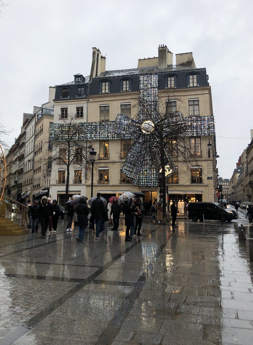 People standing on a wet city street in front of a building decorated with a large Christmas bow and ribbon installation.