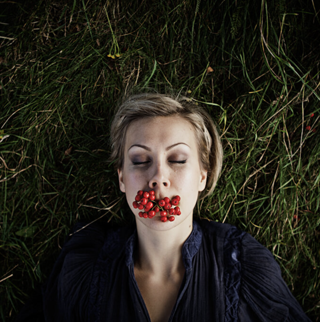 A woman with closed eyes lying on grass, with red berries over her mouth.