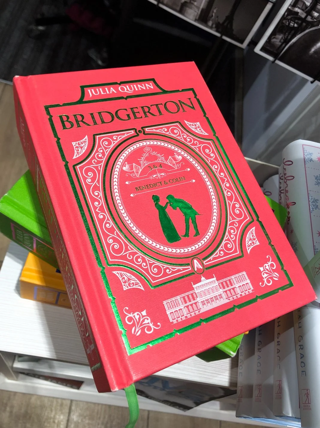 A red book titled 'Bridgerton' by Julia Quinn with green and white decorative accents, featuring an illustration of a man and woman holding hands, and a portrait of a building at the bottom.