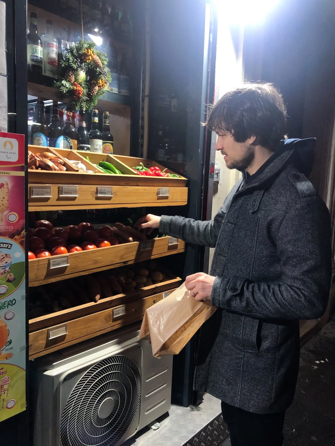 A man shopping for vegetables in a grocery store, selecting onions from a wooden display shelf with other vegetables around him.