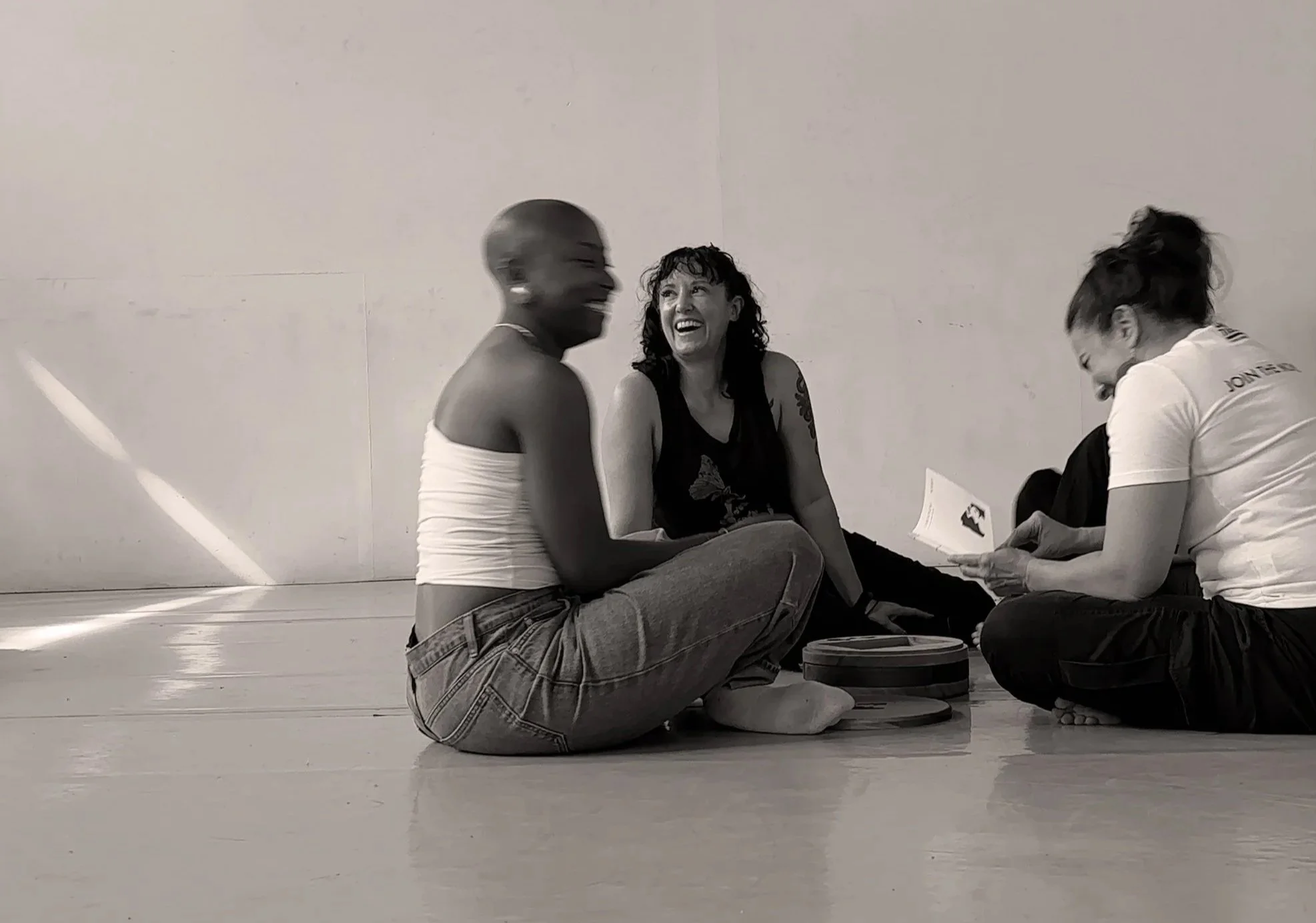 Three women sitting on the floor and talking, two appear to be laughing, in a plain room with a light-colored wall.