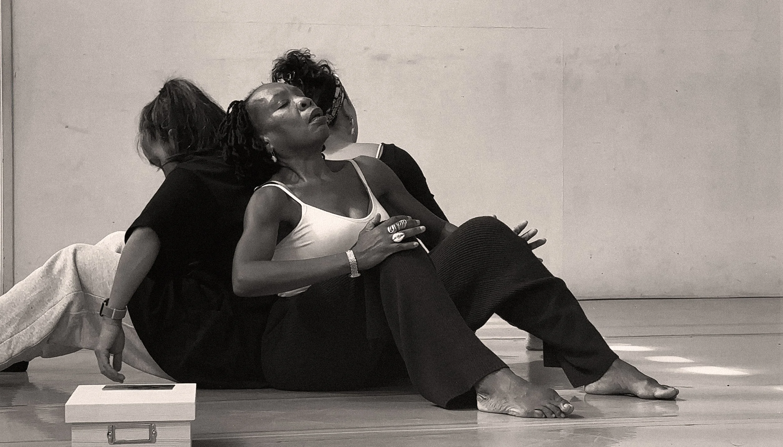 Three women sitting on the floor, two of them leaning against a wall, with one woman in the foreground with her eyes closed and hands over her stomach.