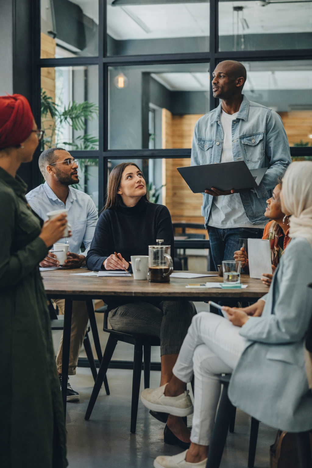 A diverse group of people participating in a meeting in a modern office space. One man is standing and speaking while holding a tablet, and others are seated at the table listening attentively, with some taking notes or holding coffee cups.