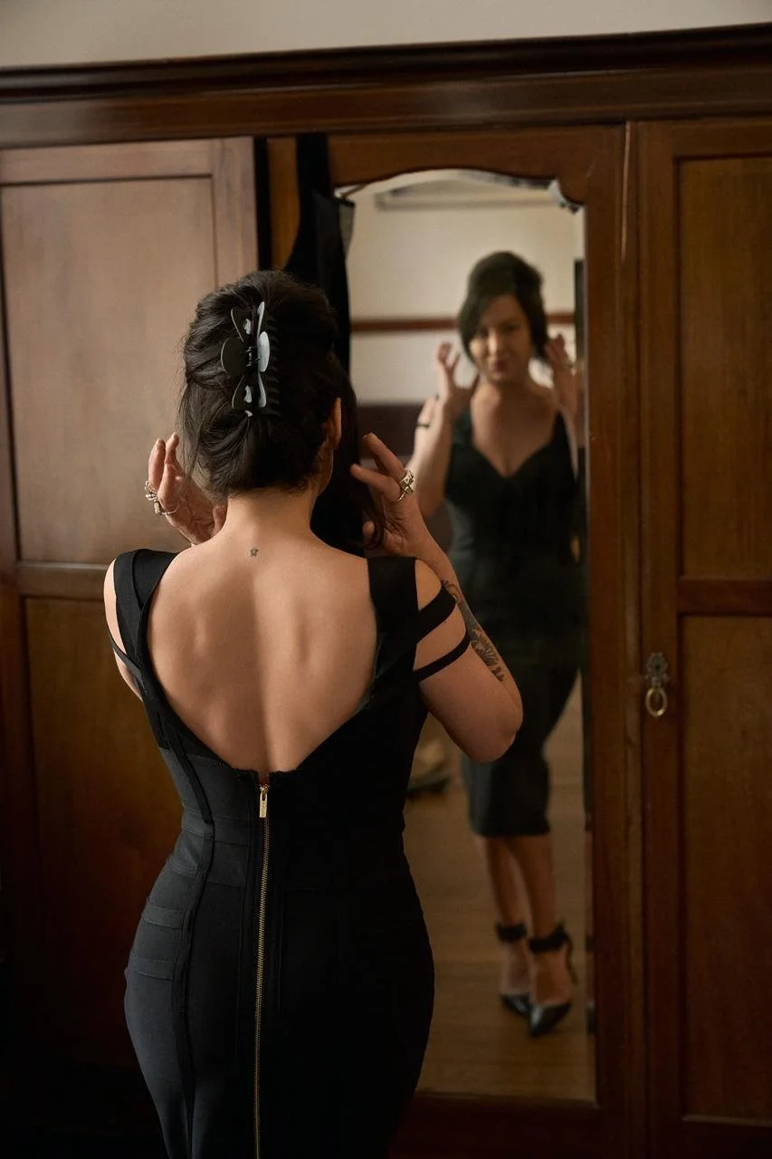 A woman looking at herself in a mirror, wearing a black dress with open back and showing tattoos on her arms and back.