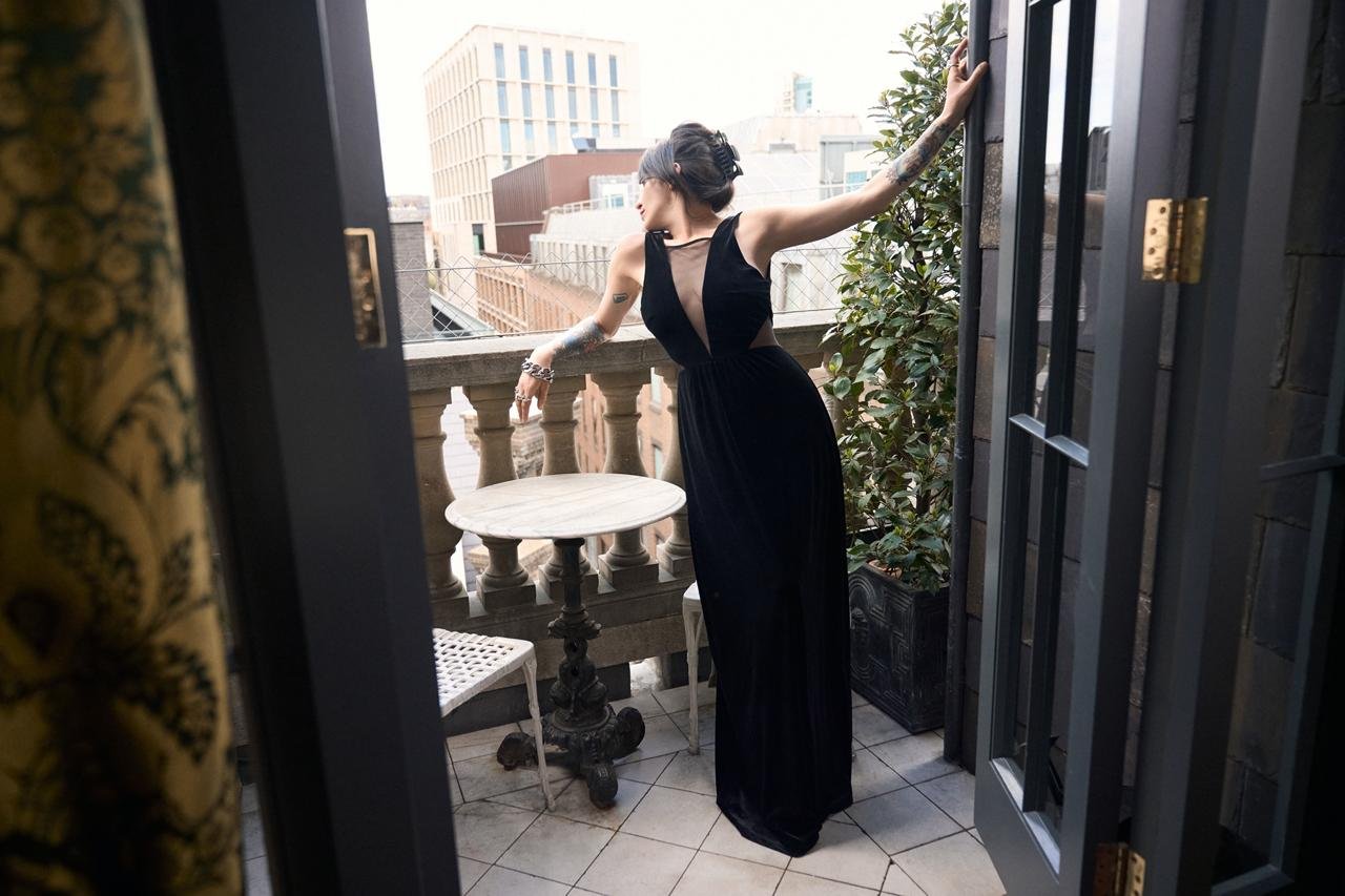 Woman in a black dress standing on a balcony, looking away, with her arm extended and resting on the balcony railing, surrounded by urban buildings and greenery.