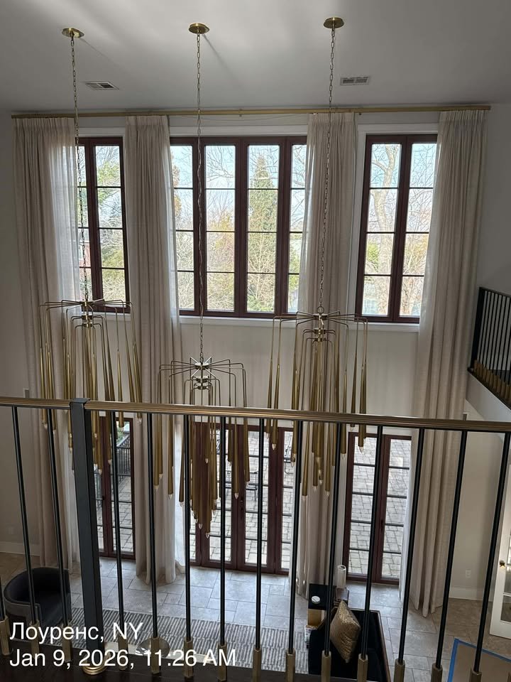 A view of a two-story living room with large windows, cream curtains, a hanging gold chandelier, a balcony railing, and a sitting area with a couch and coffee table.